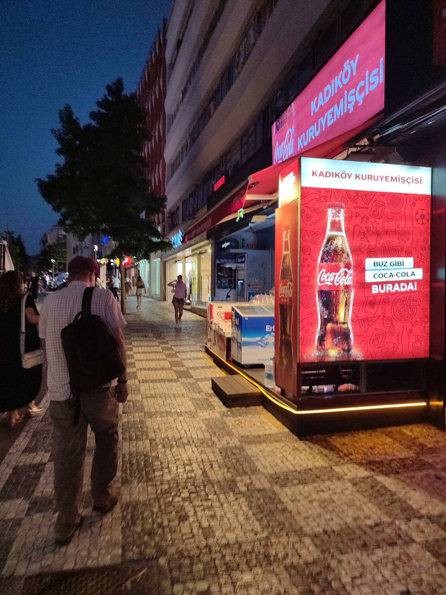 The image depicts a street scene at dusk in Kadıköy, Turkey. In the foreground, two individuals, appearing to be a couple, walk away from the camera on a patterned cobblestone sidewalk. The man in front, wearing a plaid shirt and backpack, and the woman to his left, with a white purse, are walking towards a brightly lit Coca-Cola stand. The stand features a large digital display showcasing a bottle of Coca-Cola and text in Turkish, including "BUZ GİBİ COCA-COLA BURADA!" (Ice-cold Coca-Cola is here!). Above the stand, a larger sign reads "KADIKÖY KURUYEMİŞÇİSİ" (Kadıköy Nut Store) in blue lettering on a pink background. To the left of the Coca-Cola stand, a blue and white freezer is visible. The background reveals a multi-story building with shops, lit windows, and trees. Other pedestrians are seen further down the street. The overall atmosphere suggests a commercial street in a Turkish city during evening hours.