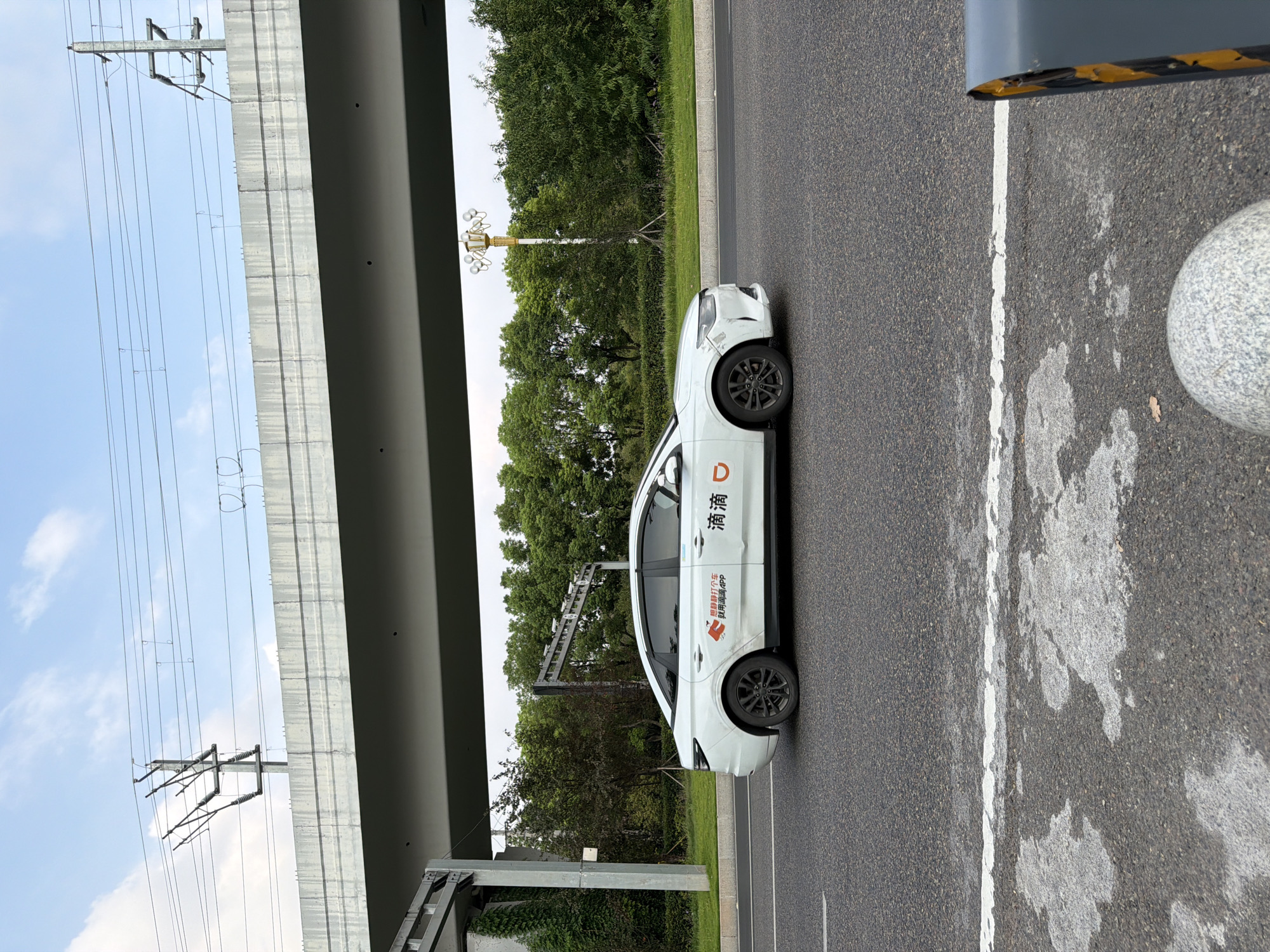 A white sedan, identified as a Didi ride-sharing vehicle by the orange logo and Chinese text on its side, drives on a paved road. The vehicle displays some damage on its front bumper. In the background, a concrete overpass with power lines stretches across the frame, supported by pillars. Lush green trees line the roadside behind the car. The sky above is partly cloudy, with a blue expanse visible. The scene appears to be set in an urban environment, likely Jiangjin, China, given the presence of the Didi branding and infrastructure. The road is asphalt with a white line marking.
