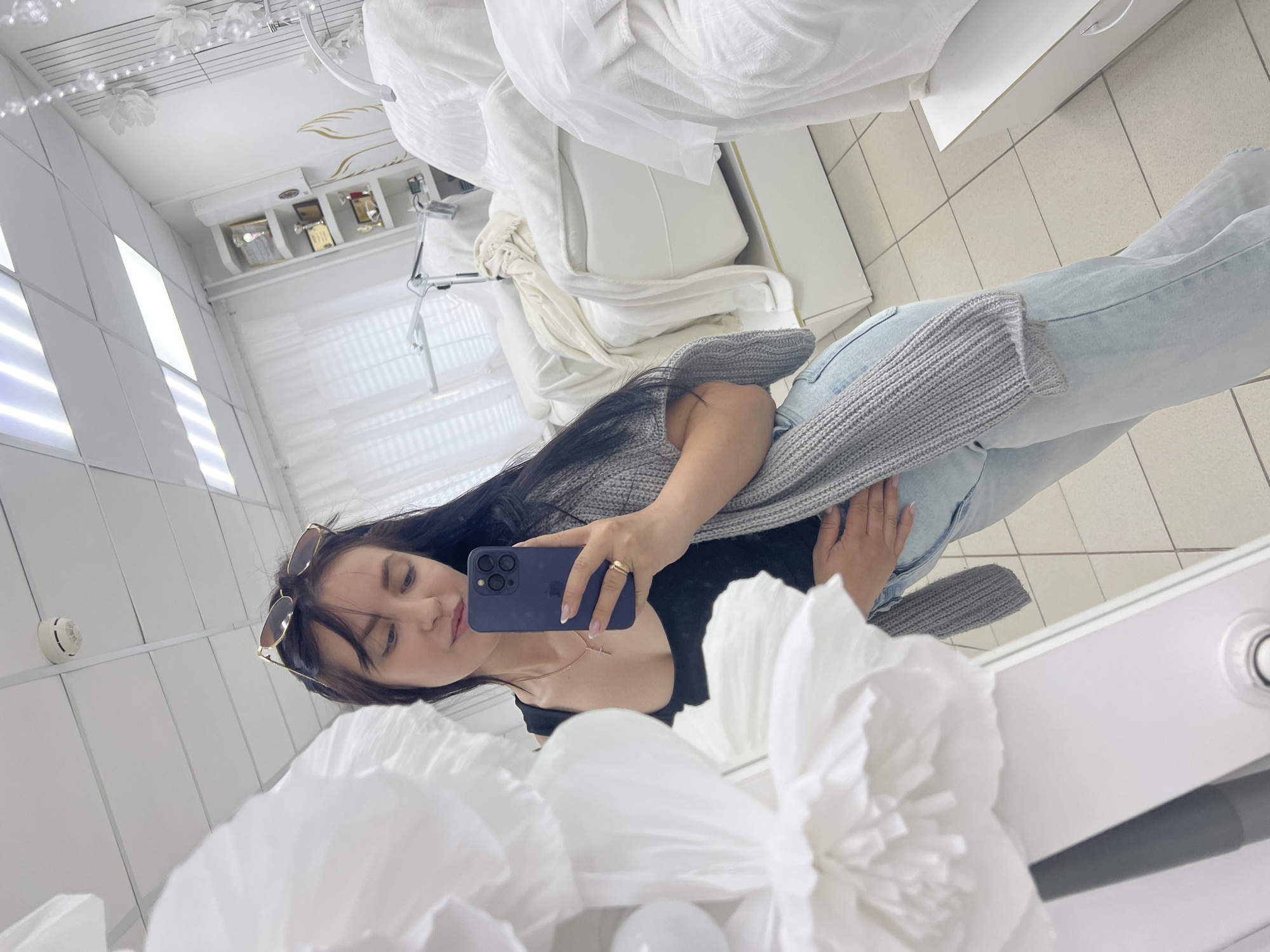 In the city of Chaikovsky, Russia, a woman with dark hair, wearing sunglasses on her head, a black top, a grey cardigan, and light blue jeans, stands in a room with white tiled floors and white walls. She is holding a dark blue smartphone in her right hand and taking a selfie in a mirror. A large white paper flower is in the foreground to the left of the frame. In the background, several white massage beds are visible, along with shelves containing framed certificates and trophies. The room appears to be a beauty salon or spa.