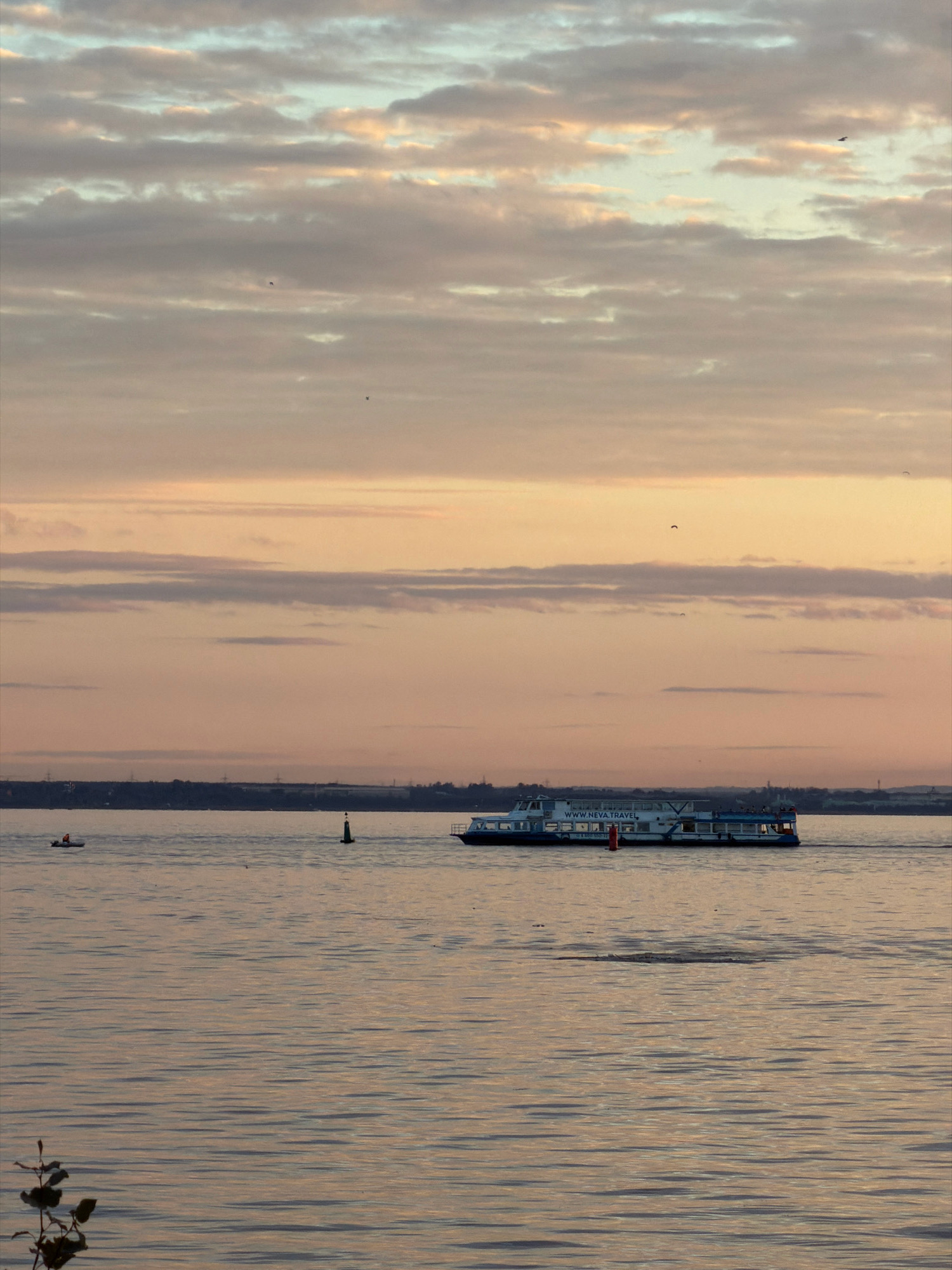 The image depicts a serene sunset over the Neva River in Saint Petersburg, Russia. The sky is a canvas of soft, pastel hues, with bands of pale orange, pink, and light blue clouds. A few small birds are visible in the distance. In the middle ground, a white passenger boat with blue trim cruises across the calm water. The boat has "WWW.NEVA.TRAVEL" written on its side. To the left of the boat, a small orange buoy bobs on the surface. Further to the left, a small motorboat with a single occupant wearing an orange life jacket is also visible. The far bank of the river is lined with a dark silhouette of trees and structures, possibly industrial, and dotted with distant lights. The foreground features the rippled surface of the Neva, reflecting the muted colors of the sky. A single, bare branch with a few leaves extends into the lower left corner of the frame.