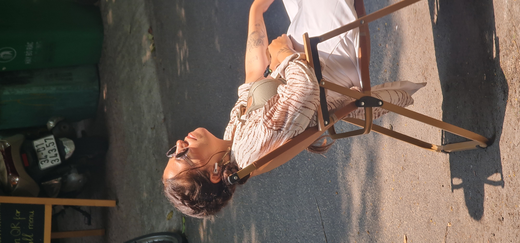 An adult female sits in a tan folding chair in an urban location. She wears sunglasses, a light-colored tiger stripe patterned shirt, and a light-colored strapless top. She has tattoos on her left arm. The chair casts a shadow on the pavement. In the background are green trash bins and a parked moped with license plate 34-01 343.57. A small wooden sign with the words "Scan QR for full menu" is partially visible. This appears to be Đà Nẵng, Vietnam.