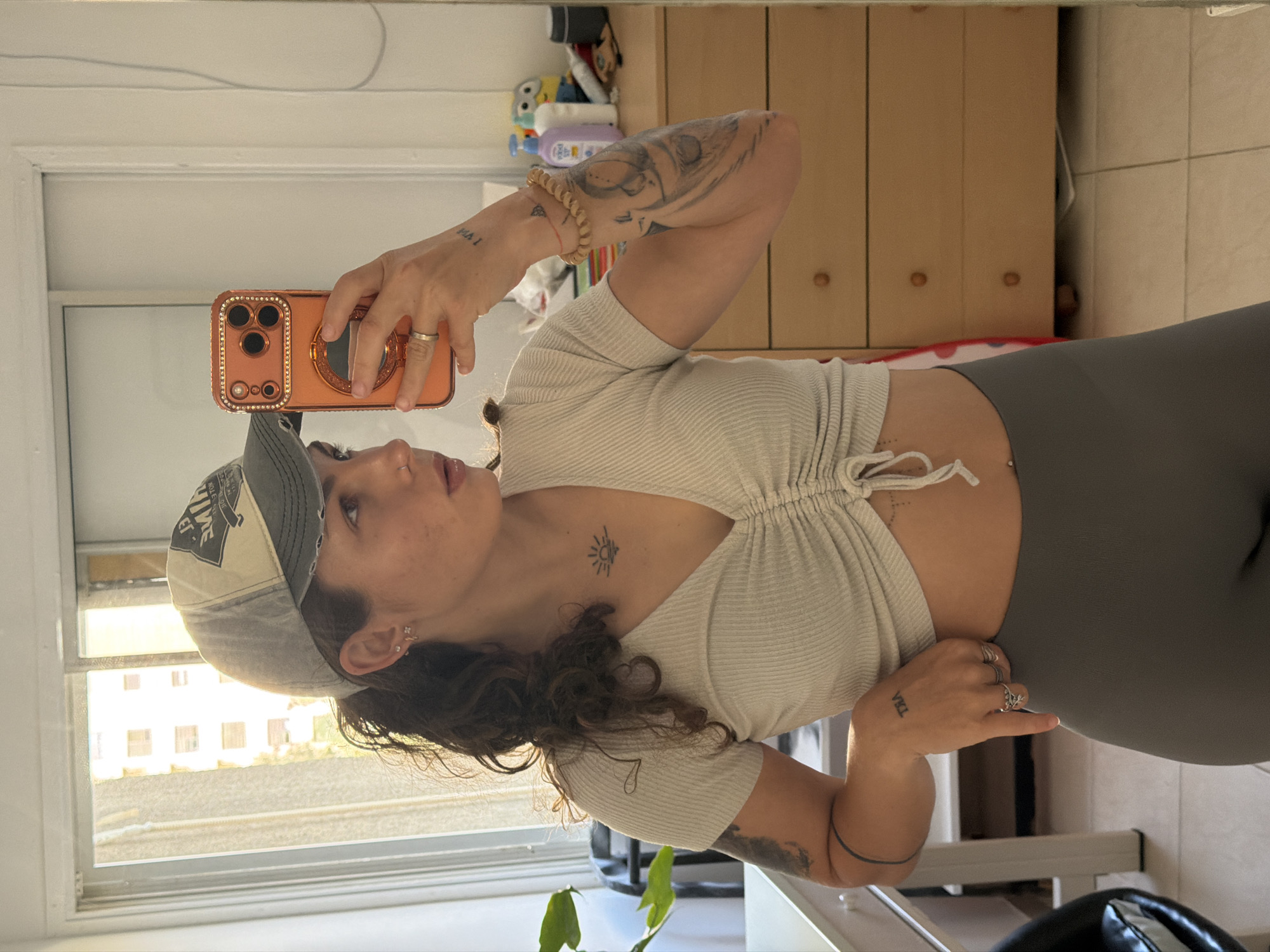 A young woman is captured in a mirror selfie, turned sideways and looking towards her phone. She has dark, curly hair, multiple tattoos, and is wearing a light-colored ribbed crop top with a drawstring front, dark gray leggings, and a gray and off-white baseball cap. The cap features text, with "NATIVE" partially visible.
Her ornate, rose gold phone case, adorned with rhinestones and a large ring holder, is held up to take the picture. Visible tattoos include a large animal head design on her left forearm, a thin circular band on her right arm, tiny dots on her lower abdomen, and a small sun with rays on her upper chest/collarbone. Text "SHA I" (or similar, inverted) is tattooed on her left wrist. She wears several rings on her fingers, including one with a leafy design on her right hand, and small earrings. A navel piercing is also visible.
The setting appears to be an indoor room, likely in Kiryat Bialik, Israel, with a large mirror reflecting the scene. In the background, a light-colored wooden cabinet with decorative knobs, and various small items and toys, can be seen. A window to the left reveals bright daylight and multi-story residential buildings outside, suggesting a sunny daytime. Part of a green plant is visible in the lower left corner. The floor is tiled.