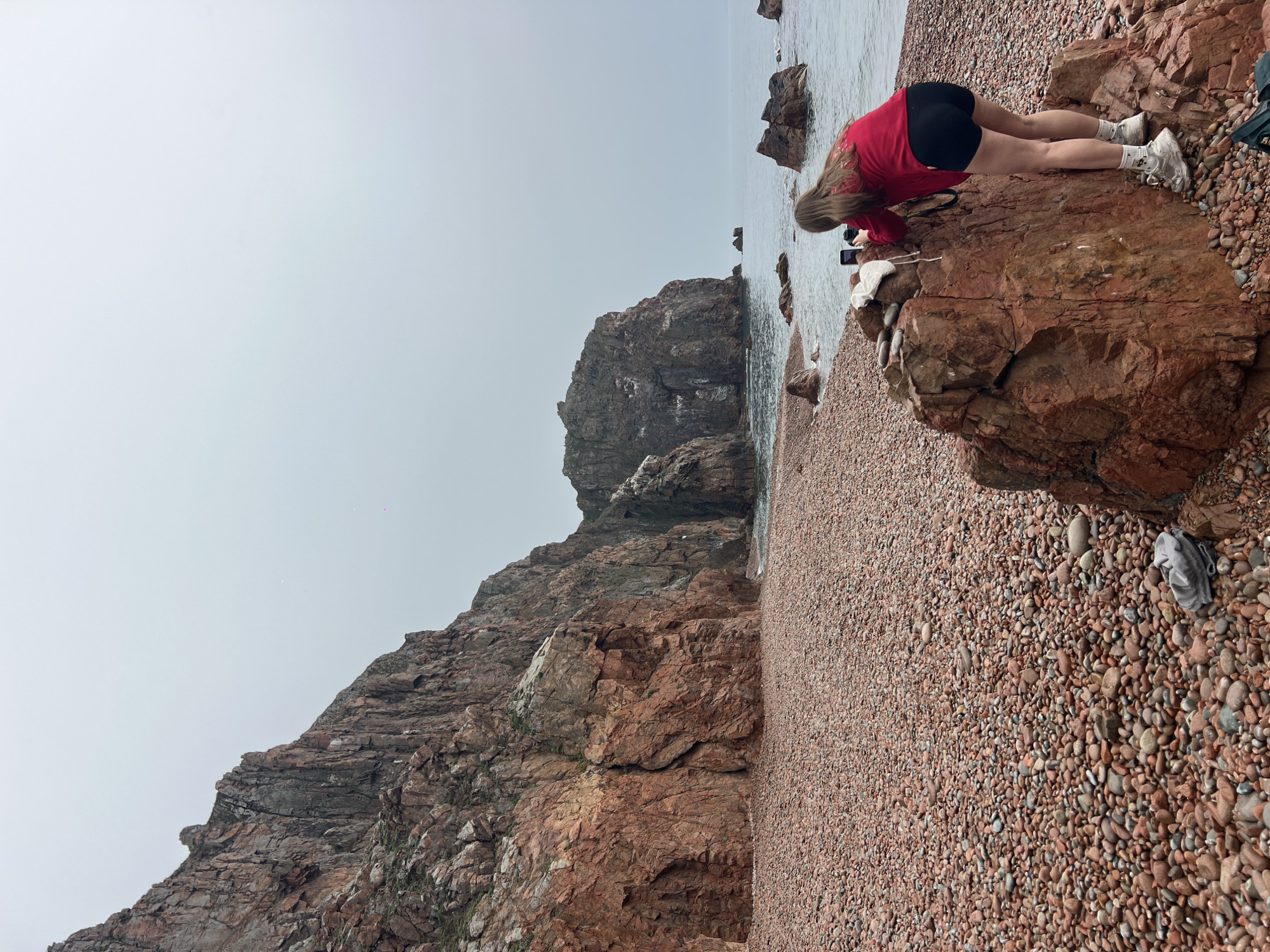 The image depicts a rocky beach in Russkiy, Russia. A woman wearing a red shirt and black shorts is leaning over a large rock near the water's edge, seemingly using a phone to take a picture. The beach is covered in small, reddish-brown pebbles. To the left, a steep, rocky cliff rises up. The sky is overcast, creating a muted color palette.