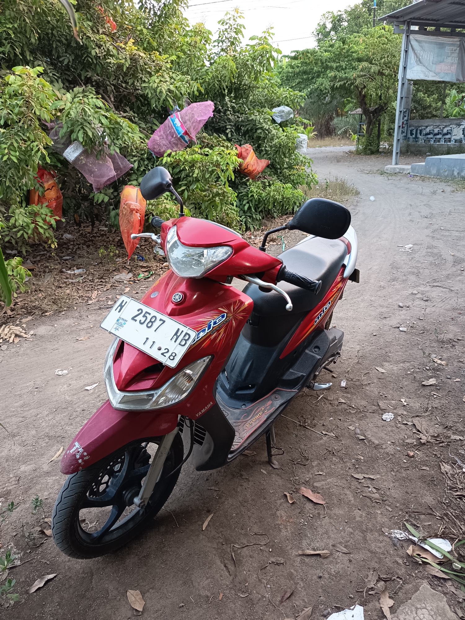 A red Yamaha Mio automatic scooter is parked on a dirt road in a semi-rural area. The scooter's license plate reads "H 2587 NB" with "11.28" below it. The scooter has "Mio" and "Sporty" graphics on its side. In the background, there are trees with various items, possibly plastic bags or fabric, hanging from them. Some of these items are red and pink. A simple structure with a metal roof is visible to the right. The ground is dry and covered with dirt, leaves, and some scattered debris. The lighting suggests it is daytime. The location is identified as Krampilan, Indonesia, based on the context provided.