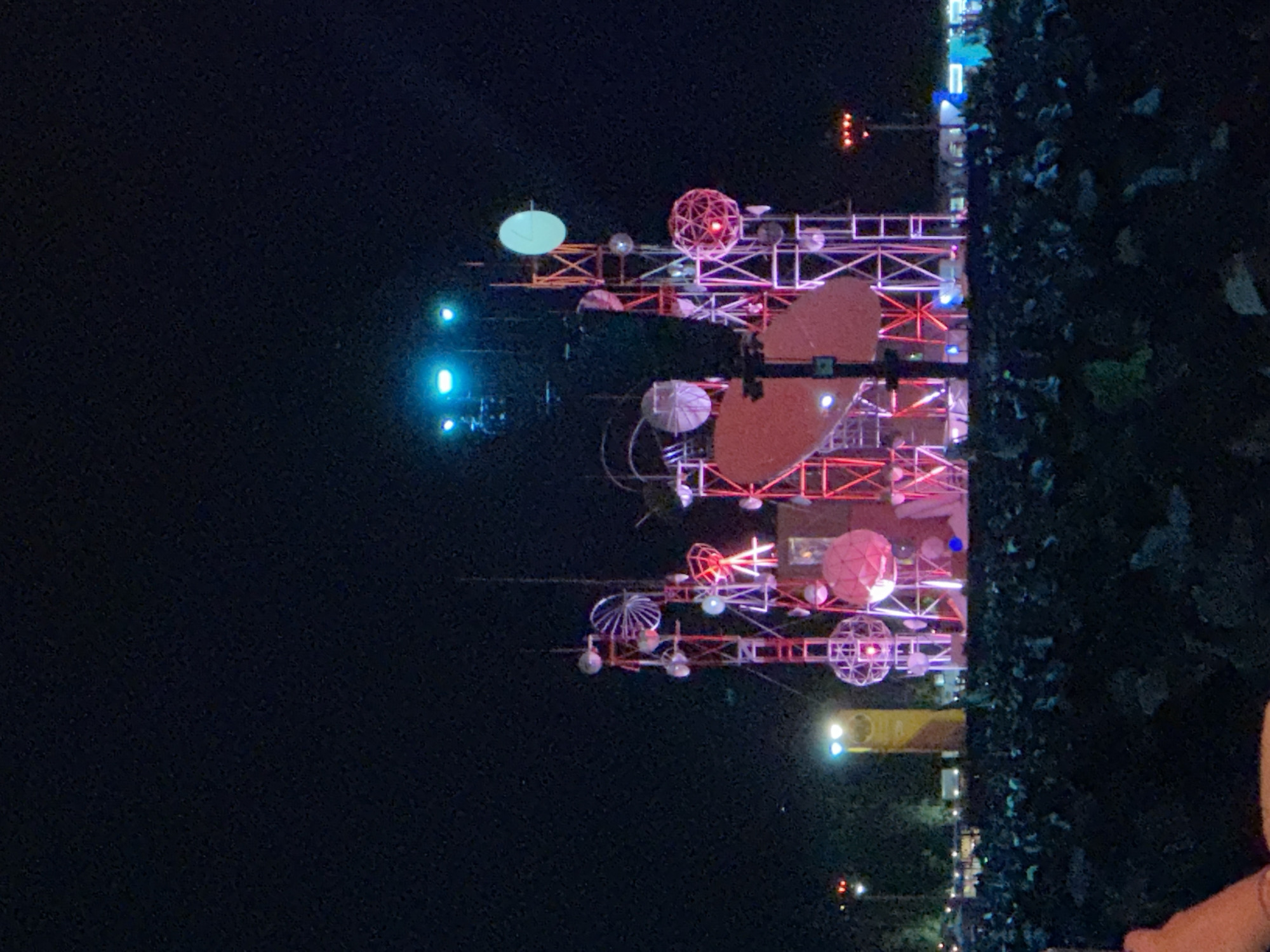 At night in Indio, California, a temporary illuminated structure stands, featuring spheres, discs, and lattice elements, some adorned with lights. The structure, partially obscured by foliage on the right, is built of metal scaffolding and other materials.