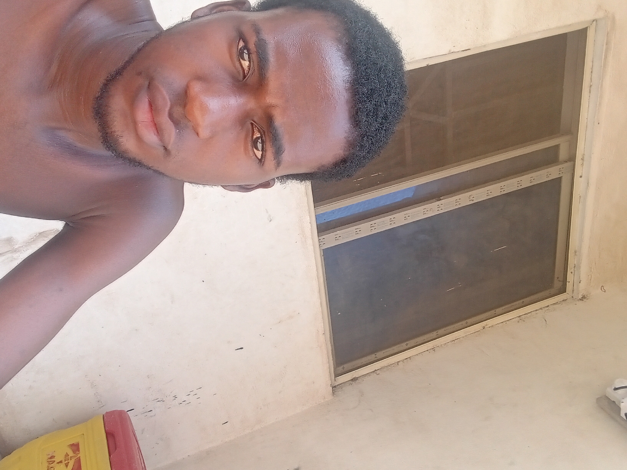 A selfie photograph depicts a shirtless young man with dark skin and short, curly black hair. He is positioned towards the left side of the frame, his face close to the camera, and his gaze directed towards the lens.
To the right of the subject, a window is visible, consisting of two panes. The window frame appears to be a light color.
The background is a wall, painted in a light color. In the bottom left corner of the frame, part of a yellow and red object is visible.
Based on the style of building, the type of window and the subject's appearance, the location could potentially be Lokoja, Nigeria, though there is no definitive proof of this in the image.