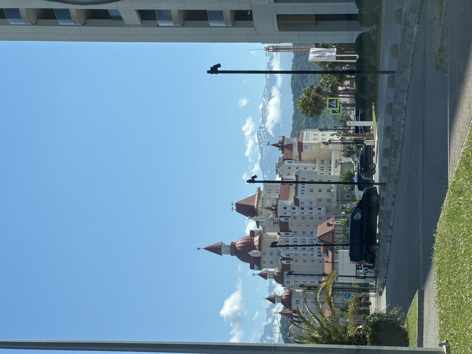This image shows a view of the Radisson Blu Resort & Congress Centre in Sirius, Russia. The resort's distinctive castle-like architecture with red-tiled roofs and multiple turrets is prominently featured. In the background, snow-capped mountains rise above a layer of clouds, suggesting a mountainous region. In the foreground, a street with a black van and other vehicles is visible, along with palm trees and green landscaping. A modern, light-colored building with balconies is on the right side of the frame, partially obscuring the view of the resort. The sky is clear and blue with scattered white clouds. Traffic signs, including a pedestrian crossing signal, are present along the road. The overall scene depicts a bright, sunny day in a resort town.