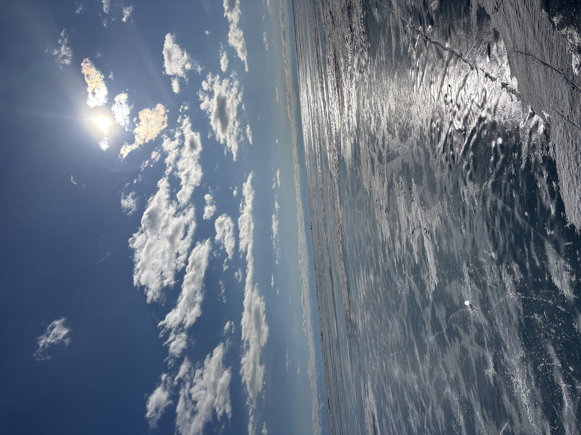 The scene depicts the frozen surface of Lake Baikal, off the coast of Listvyanka, Russia, under a clear blue sky with scattered white cumulus clouds. The sun is positioned in the upper right quadrant, emitting a bright glare and creating iridescent hues on nearby clouds. A distant range of snow-capped mountains or hills defines the horizon. The ice covering the foreground and mid-ground is a vast, semi-translucent blue expanse, marked by numerous fine white linear cracks and patches of light snow or frost. A strong band of specular sunlight reflection extends across the ice from the mid-ground towards the right. Several small, indistinct human figures are visible in the far distance on the ice, appearing to move. A single, small, white, circular object, consistent with a puck or ball, rests on the ice in the lower-left-mid section of the frame.