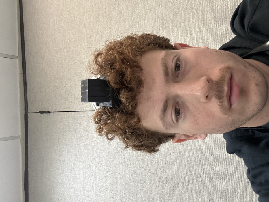 A close-up, frontal image depicts a young adult male with curly, reddish-brown hair and light skin. A black tefillin, specifically the *shel rosh* (head-tefillin) component consisting of stacked rectangular blocks with visible wiring, rests on top of his head, embedded in his hair. His brown eyes are directed downwards, and he has a light mustache. He wears a dark-colored hooded garment, with a dark lanyard or strap visible near his neck. The background features a light-colored, textured wall, likely an office partition, which meets a drop ceiling composed of light-colored acoustical tiles and a visible grid. A thin, black vertical wire is visible against the wall in the upper left portion of the frame. The image depicts an indoor environment. No specific visual identifiers within the image allow for the determination of its precise location within Fairfield, United States of America.