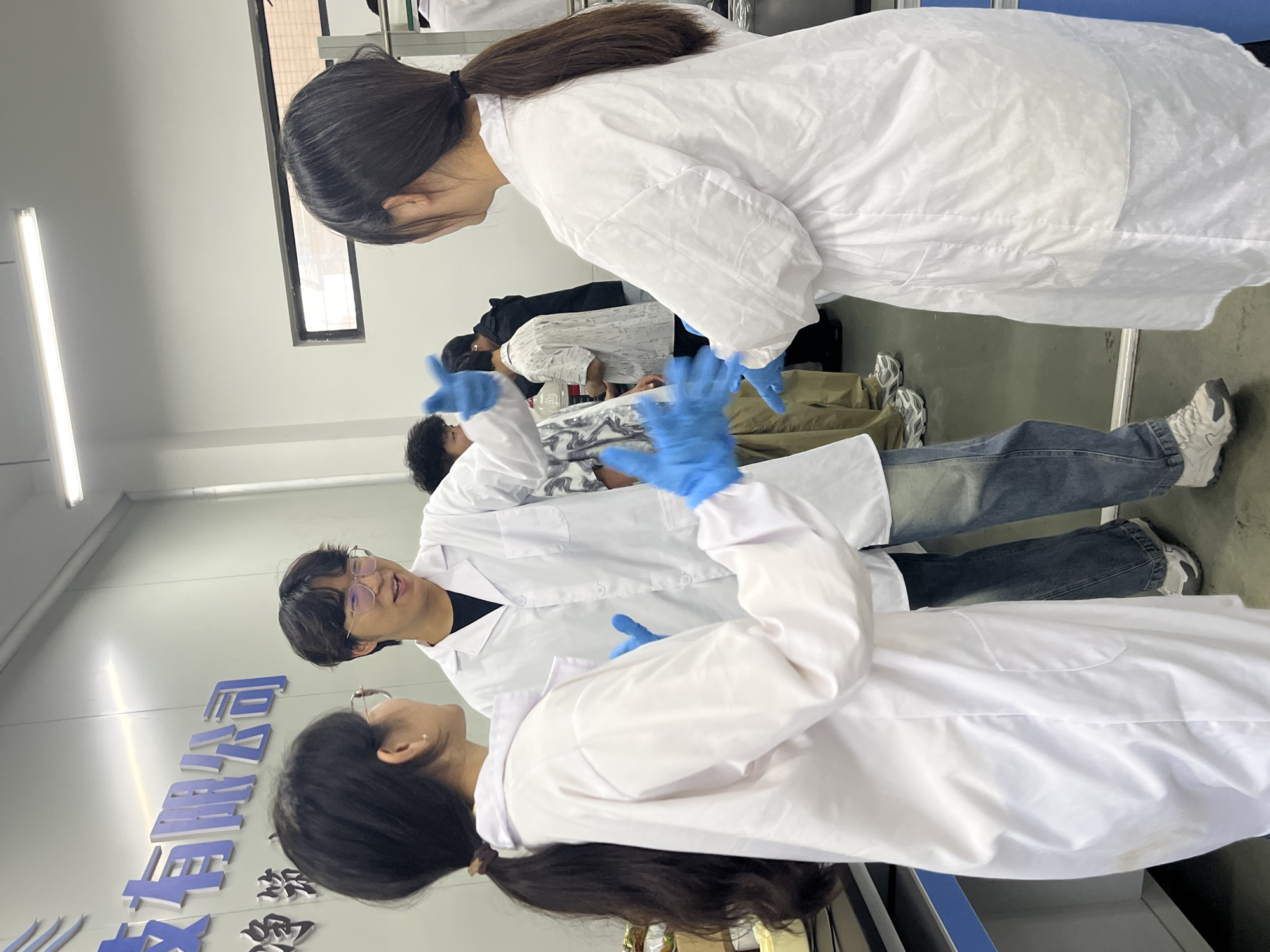 The image shows a group of people in what appears to be a laboratory setting, possibly in Jiangjin, China. Several individuals are wearing white lab coats and blue gloves. One person is wearing glasses. The setting is indoors, with fluorescent lighting and an indistinct background. There is Chinese writing on a wall. The image shows people interacting and engaged in discussion. The exact purpose of their activity is not clear from the image.