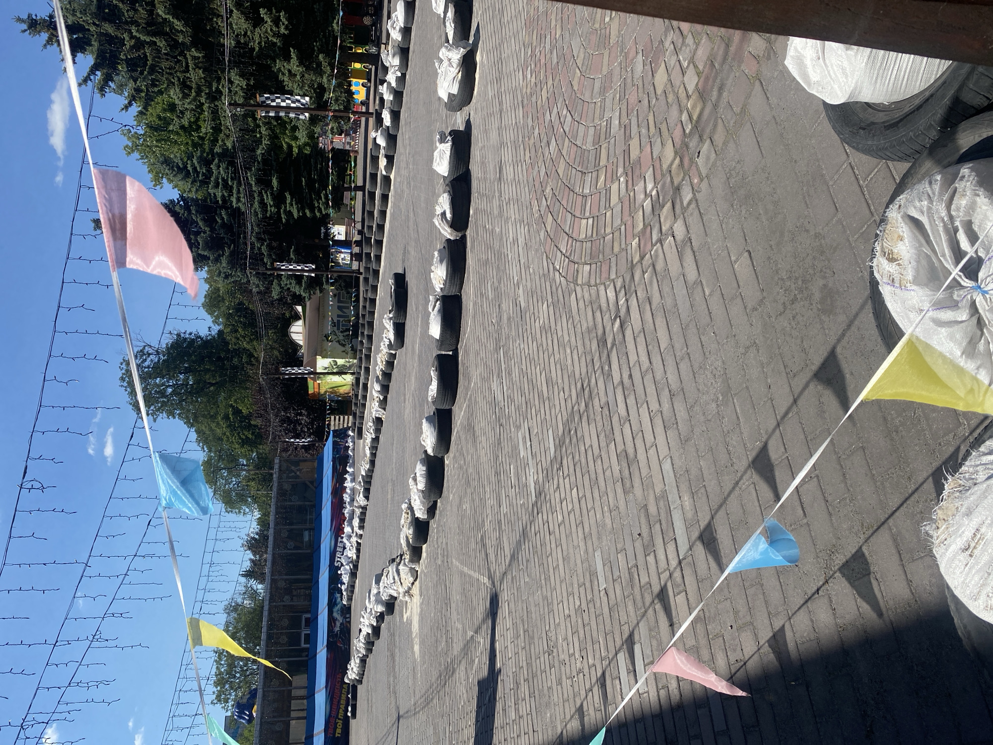 The image shows an outdoor go-kart track in Kamianske, Ukraine. The track is paved with brick pavers and lined with tires filled with white sacks, serving as barriers. Festive pennant banners in pink, blue, and yellow are strung across the track, along with string lights. In the background, there is a blue building with text that appears to be in Ukrainian, along with trees and other structures that suggest an amusement or recreational area. Several checkered flags are visible on poles. The scene is set on a bright, sunny day.