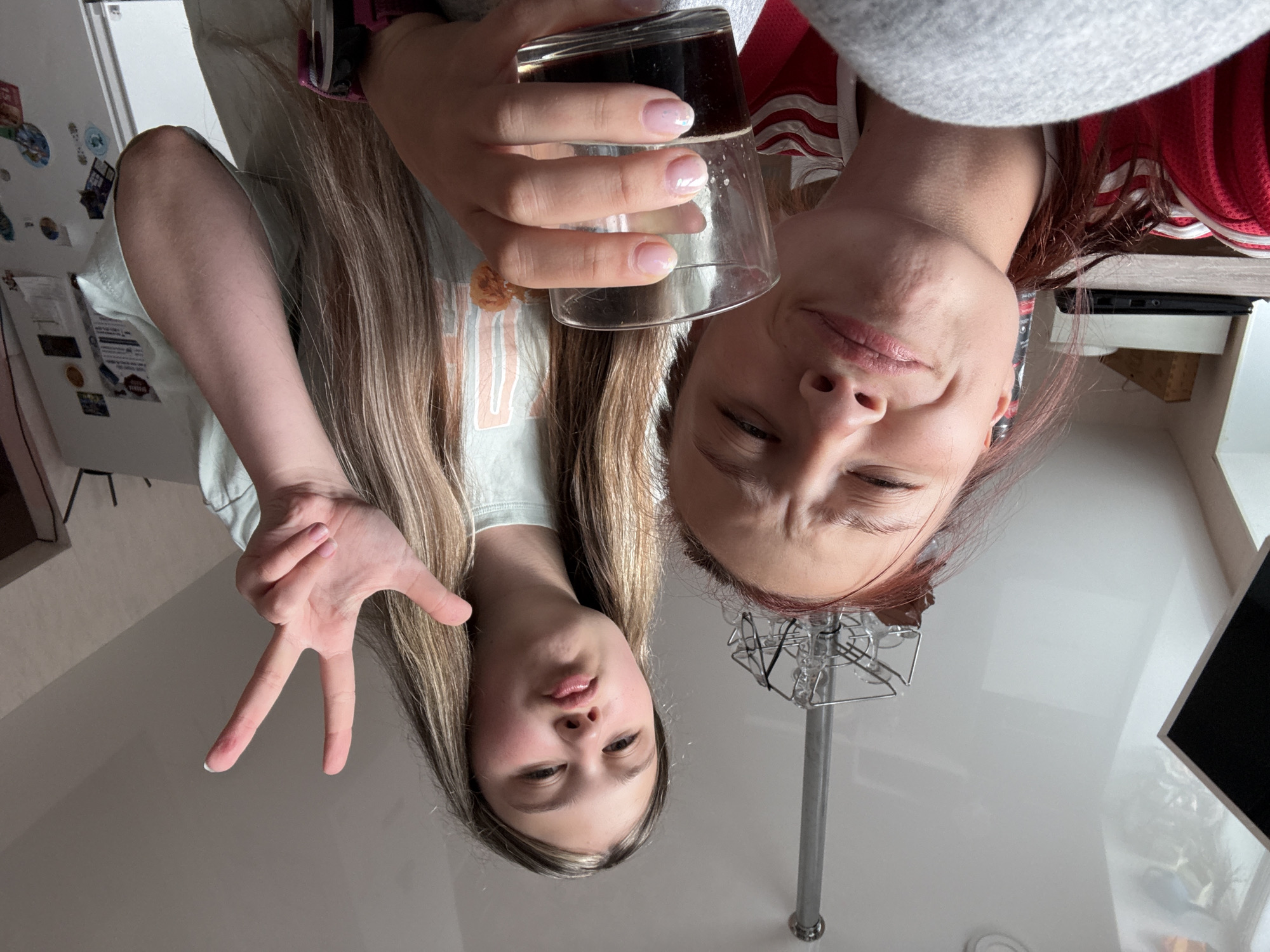 Two women are facing downward towards the viewer. The woman on the left is holding up two fingers in a "peace sign." The woman on the right is holding a glass of dark liquid. A metal stand extends upwards between their faces. The setting appears to be indoors, possibly a kitchen or bathroom, given the presence of a countertop and a partial view of a monitor. The location is unspecified beyond the presence of the Russian city of Прикамский.