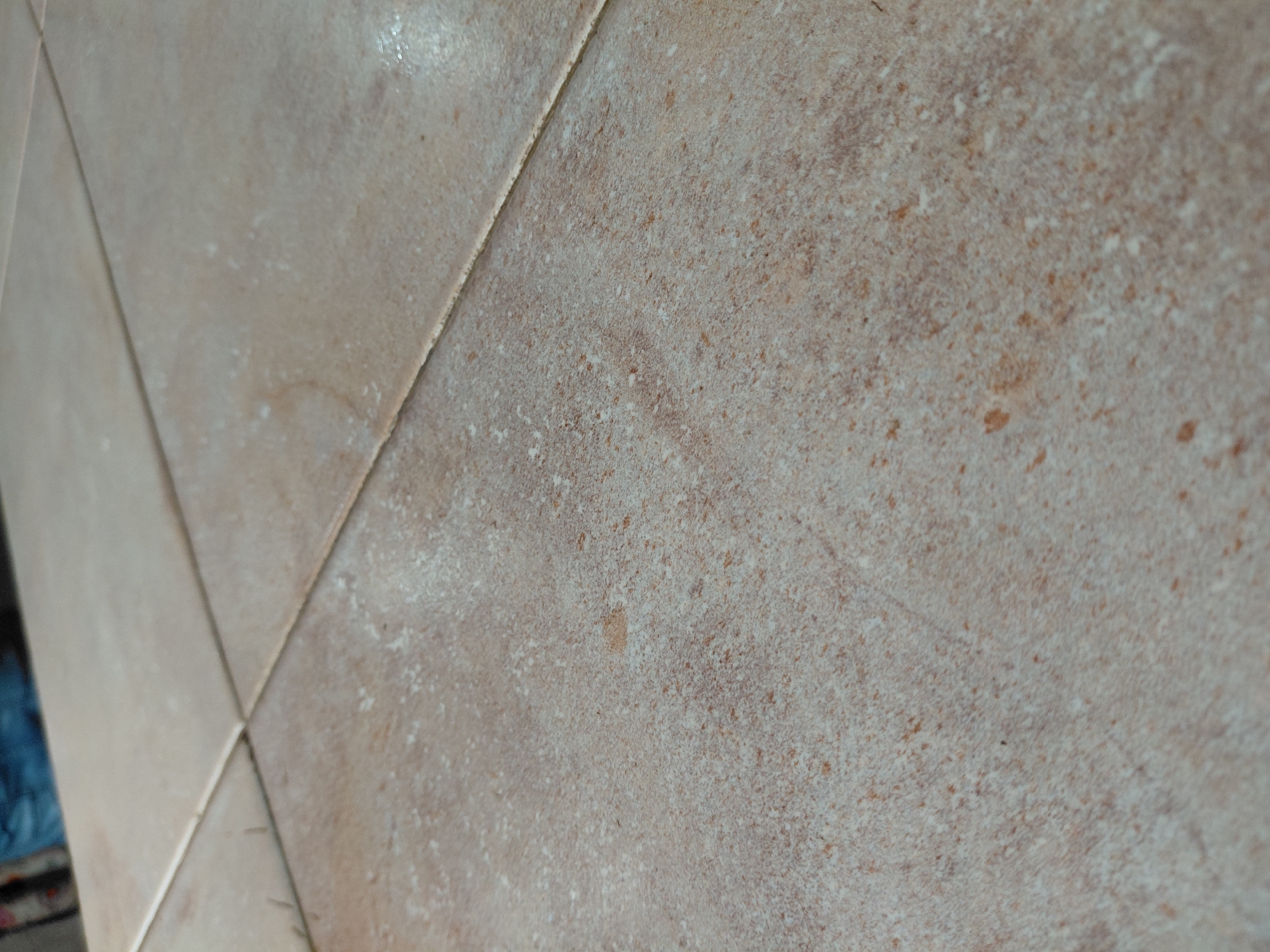The image presents a close-up, oblique view of a tiled surface, likely a wall or floor, located in Santa Ana, Venezuela. The tiles are light brown, beige, and tan, exhibiting a distinct mottled and speckled texture. This texture is characterized by numerous small, irregular white and reddish-brown flecks spread across the lighter base color.
Prominent light-colored grout lines divide the individual tiles, with one section showing a clear intersection where four tiles meet. The overall appearance suggests a textured ceramic, porcelain, or natural stone-like material. Some areas of the surface show a subtle sheen, possibly indicating light reflection or moisture. No people or specific activities are taking place. In the background of one of the cropped views, a blurred blue object is faintly visible, but its specific identity or purpose cannot be determined. There is no visible text.