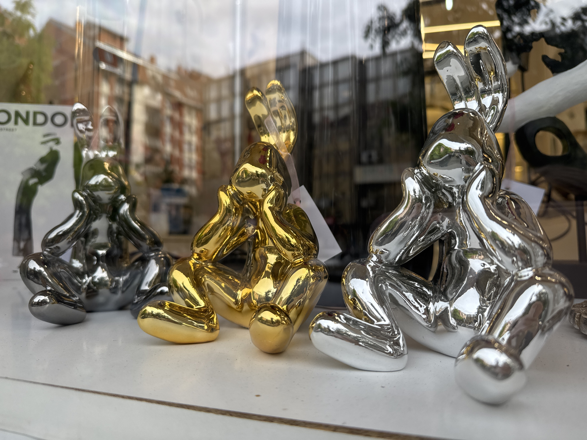 Three glossy, metallic bunny sculptures are arranged on a white surface. From left to right, they are gray, gold, and silver. Each bunny is seated with their hands on their face, seemingly in contemplation. The sculptures are viewed through a window, reflecting the outside cityscape, suggesting a store display. The reflections are blurry and feature buildings and a leafy tree. The text "ONDON" is partially visible on a sign to the left of the gray bunny. While I am unable to identify the location of the city or country from the image, the prompt suggests Novi Sad, Serbia.