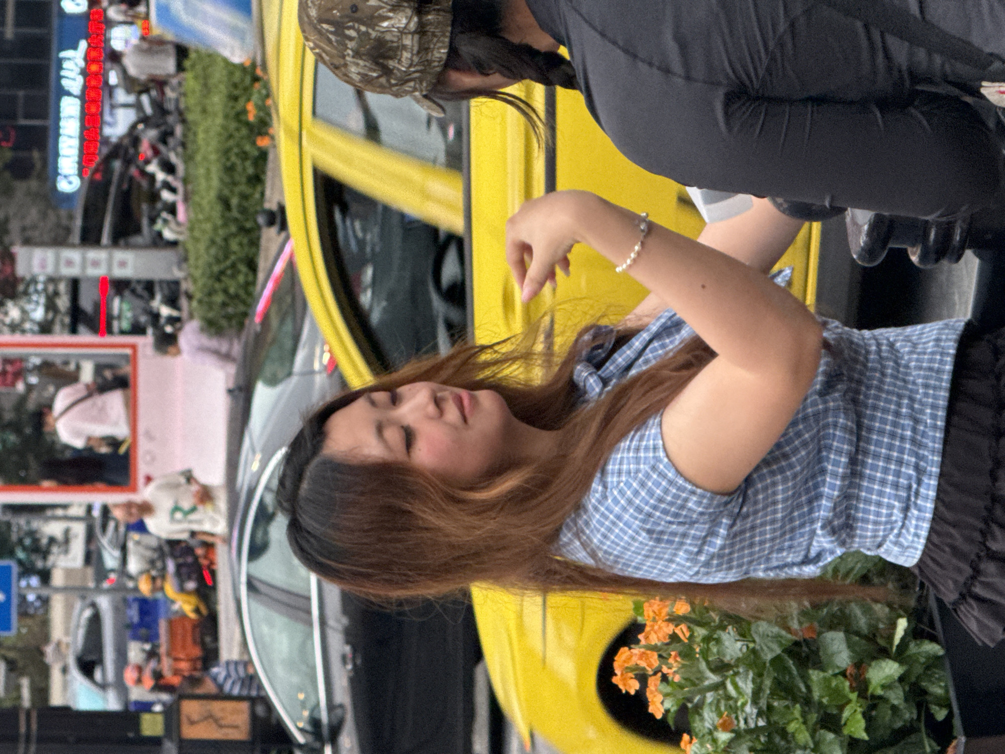 In Yuzhong District, China, a young woman with long, wavy brown hair and fair skin, wearing a blue and white plaid shirt and a black ruffled skirt, stands near a vibrant yellow car and a planter filled with orange flowers. She is looking to her right, her left hand is raised and adorned with a pearl bracelet. Her right arm is not visible. To her right, another person, seen from behind, wears a camouflage cap and a black shirt. The background depicts a busy urban street scene with various vehicles, including cars and a motorbike, and a blurred neon sign. The lighting suggests it is daytime.
