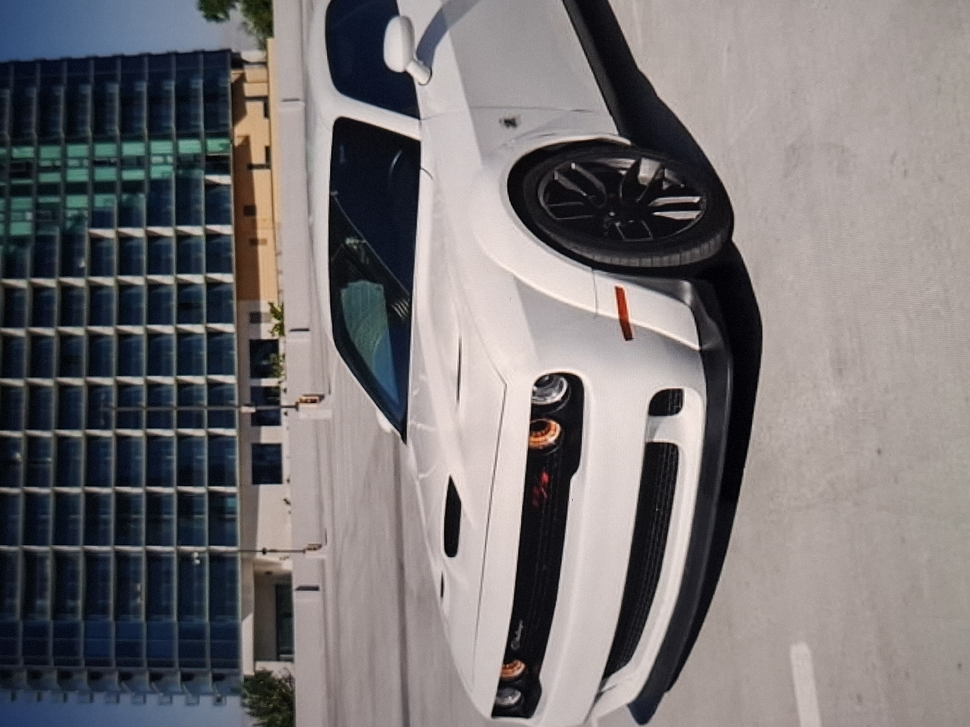The image shows a white Dodge Challenger parked in an outdoor parking lot. In the background, a modern, multi-story building with a glass facade dominates the scene. The sky is clear and blue. The car's front end, including the grille, headlights, and hood scoop, is prominently featured. The parking lot surface appears to be concrete, with some faint white lines visible. There are no people or animals in the image. This scene does not depict the city of Urus-Martan, Russia.