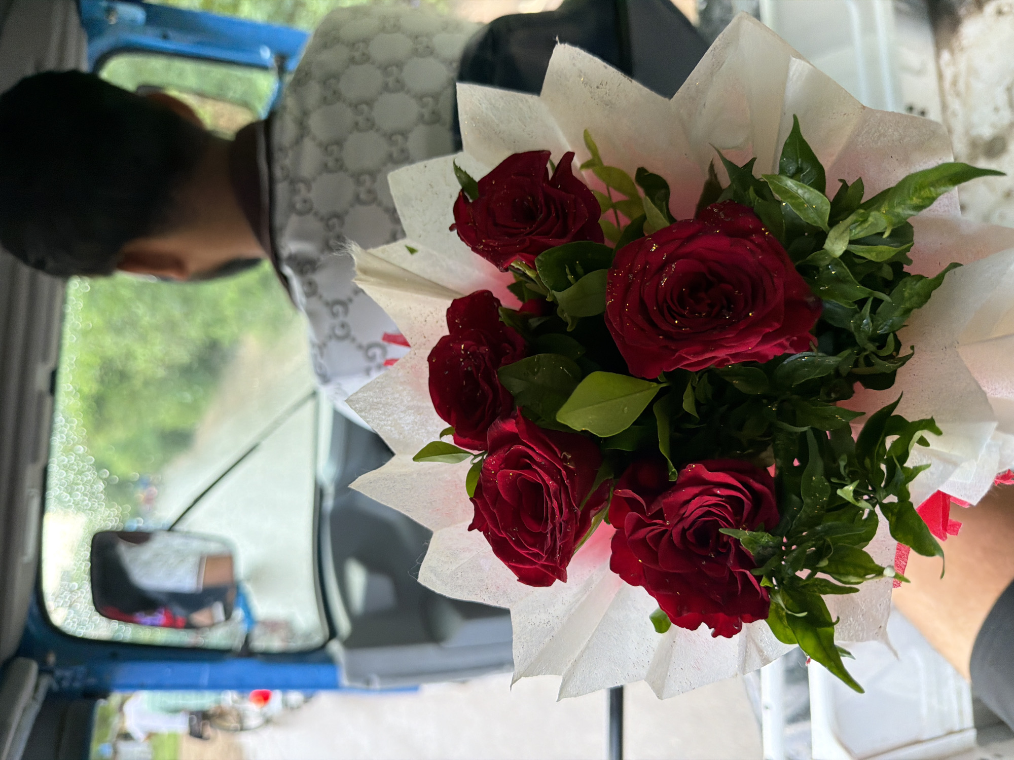 A bouquet of five red roses, with some water droplets on the petals and green leaves, is held in the foreground of the image. The bouquet is wrapped in white paper. In the background, a man wearing a white shirt with a grey pattern is driving an auto-rickshaw. Through the windshield, a road with trees and other vehicles can be seen. The scene appears to be in a city, possibly Kanpur, India, given the presence of an auto-rickshaw and the general environment. The weather seems to be wet, as indicated by the water droplets on the windshield.