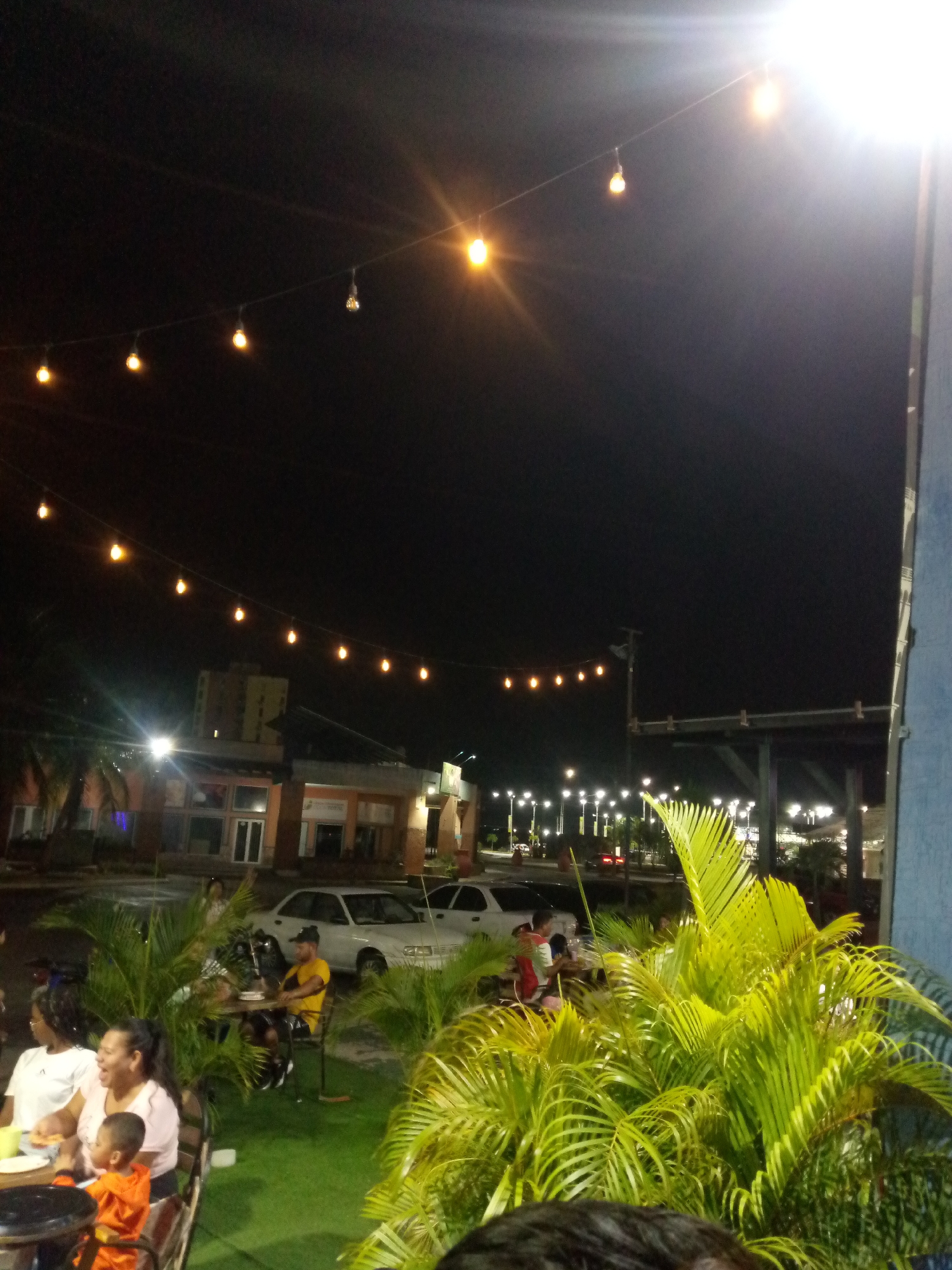 The image depicts an outdoor dining area at night. Strings of lights are strung overhead, illuminating the space. People are seated at tables, some eating, and engaging in conversation. In the background, buildings and streetlights are visible, suggesting an urban setting. Cars are parked in the distance. The specific city and area are not discernible from the visual data. Tropical plants are clustered in the foreground.