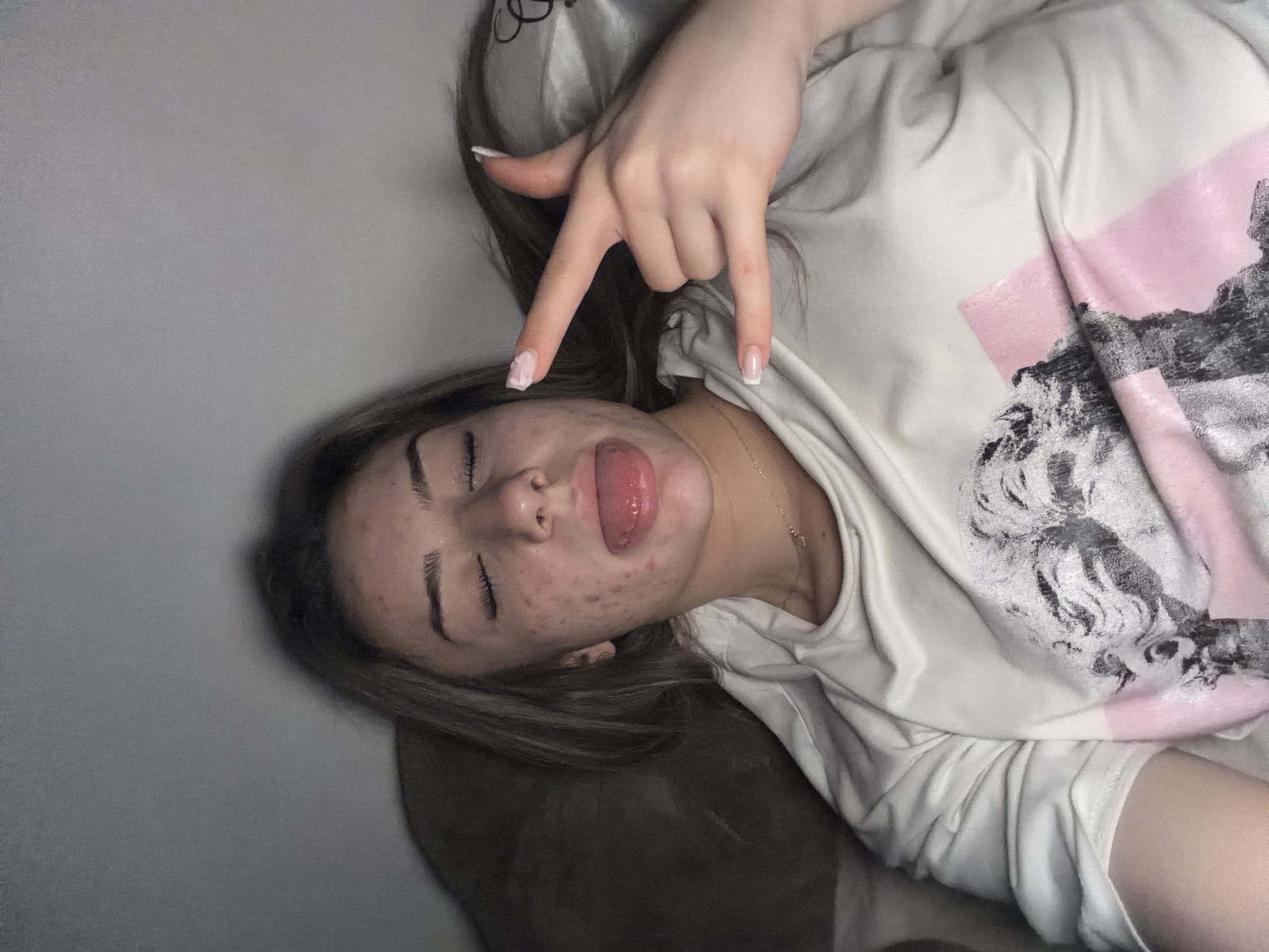 The image shows a young woman with her eyes closed, sticking out her tongue and making a rock-on hand gesture. She is lying down, possibly in bed, with her head resting on a pillow. Her face has acne. She is wearing a white t-shirt with a graphic print. The background is dark and appears to be a bedroom environment. There is no visible indication of the city of Shumen, Bulgaria.