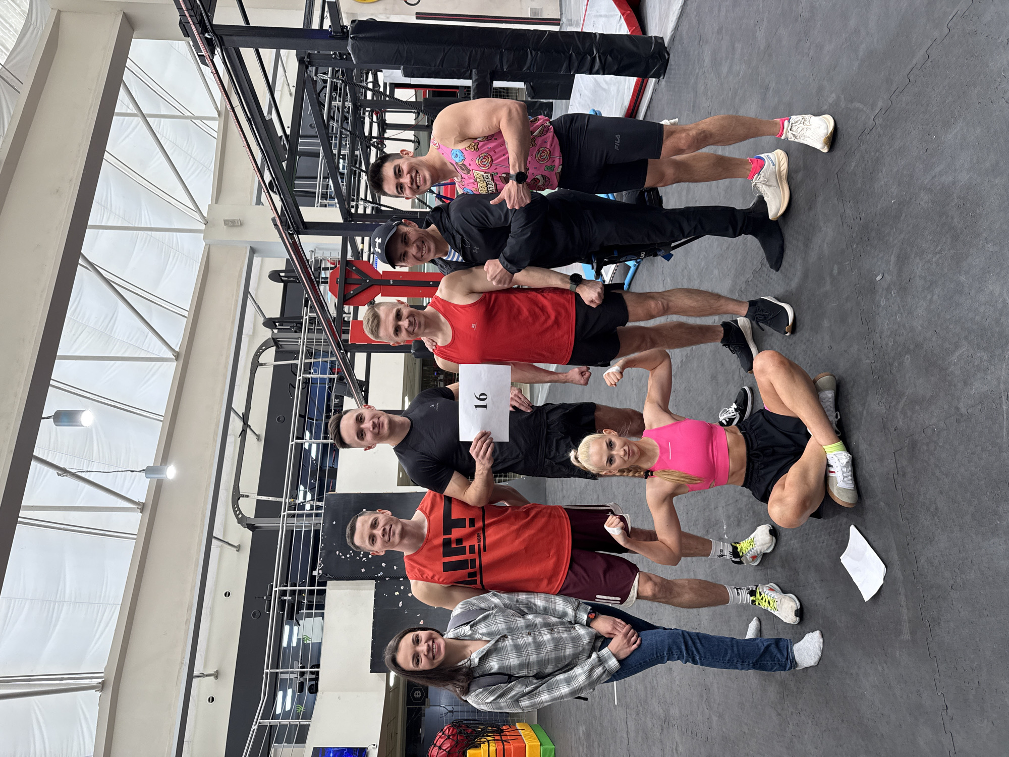 A group of six people stand in a line, possibly posing for a picture in a gym or fitness facility. The people are of varying heights and appearances, with one woman at the bottom of the line dressed in jeans and a plaid shirt, and others wearing athletic attire such as shorts, tank tops, and workout leggings. One man is holding a sign with the number "16" written on it. The background reveals gymnastic rings, pull-up bars, and other fitness equipment. The location, Kazan, Russia, is not directly visible in the image and is based on the provided request. The people are interacting by standing close together, possibly touching or leaning on each other. The floor is a dark gray, possibly made of rubber or a similar material.