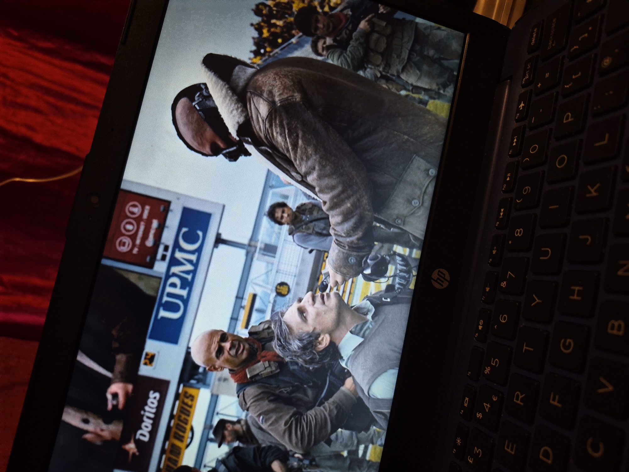 The image displays a laptop screen showing a scene from a movie. On the screen, a bald man with a red scarf is looking intensely at another man with grey hair who is being held. A man wearing a dark mask and a shearling-lined jacket, likely the antagonist, is standing behind the grey-haired man, with his hand near the man's head. Several other individuals in military-style clothing are visible in the background, along with a blurred crowd of spectators. Above the scene on the screen, there are advertisements for "Doritos" and "UPMC". The laptop itself is dark, with a visible HP logo, and its keyboard is illuminated by soft lighting, showing keys like "E", "R", "T", "Y", "U", "S", "D", "F", "G", "H", "J", "C", "V", "B", "N". The overall setting of the movie scene appears to be an outdoor public event. There is no information in the image to indicate a location in the city of Sandwell, United Kingdom.