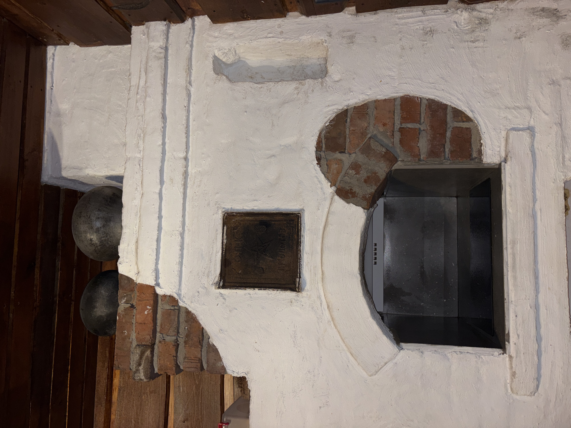A skewed, close-up shot reveals a white, ornate, plastered oven structure integrated into a wall of dark wood in a building in Perm, Russia. The oven features an arched opening framed by exposed red bricks, currently occupied by a modern, dark, rectangular appliance, likely a stove or oven insert. To the left of the opening is a square, bronze-colored plaque with possible Russian inscriptions. Two metallic spheres or globes are attached to the left of the structure, partially obscured by the wall. The plastered surfaces are roughly textured, indicative of a traditional construction method. The dark wooden wall suggests an interior setting.