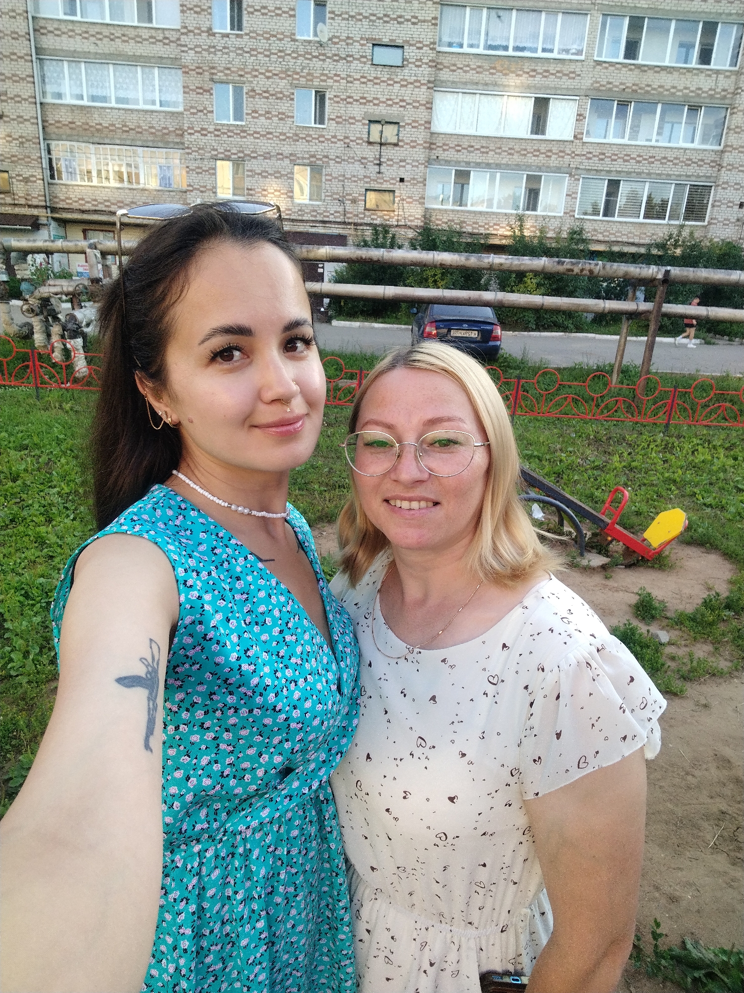 Two women are taking a selfie in an outdoor courtyard in Yanaul, Russia. The woman on the left has dark hair pulled back in a ponytail and is wearing a blue floral dress and a beaded necklace. She has a tattoo on her left arm and earrings. The woman on the right has blonde hair and is wearing glasses and a white top with a heart pattern. They are standing in front of a brick apartment building with multiple windows. Behind them, there is a green lawn, a red metal fence, a dark blue car, and playground equipment, including a seesaw. The sky is overcast.