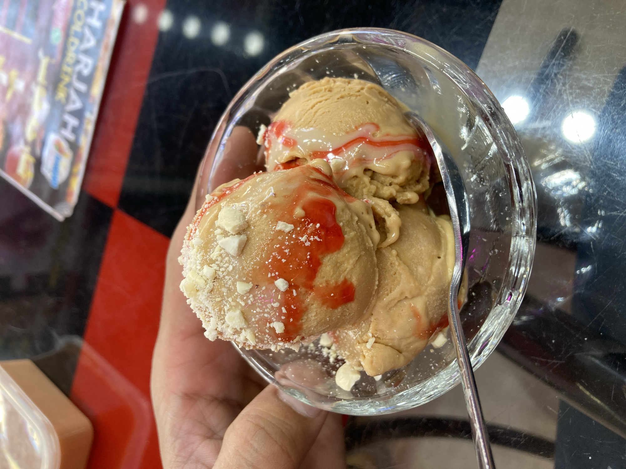 A hand holds a clear glass bowl containing three scoops of light brown ice cream. The ice cream is topped with a light drizzle of red syrup and scattered white chocolate shavings. A silver spoon rests in the bowl. A magazine with text including "COLD DRINK" and "HARJAAH" is partially visible to the left. The table has a black and red checkered pattern. This image seems to have been taken in a restaurant or cafe somewhere in Quetta City Tehsil, Pakistan.