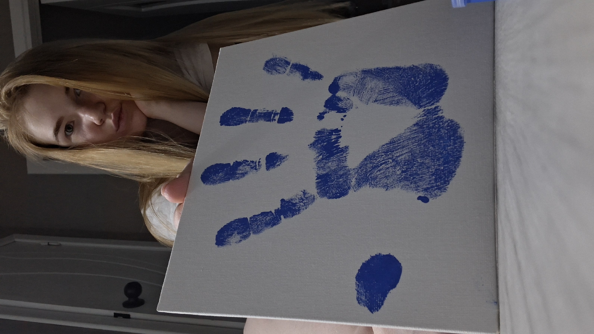 A young, light-skinned person with long blonde hair and light-colored eyes is holding a white square canvas, presumably in an indoor setting. A blue handprint is prominently displayed on the canvas. A separate blue blotch of paint is visible in the lower right corner of the canvas. The person is resting their head on their hand, looking at the viewer. A partially visible door can be seen in the background. The location is unknown and there is no indication it is in the city of Chaikovsky, Russia.