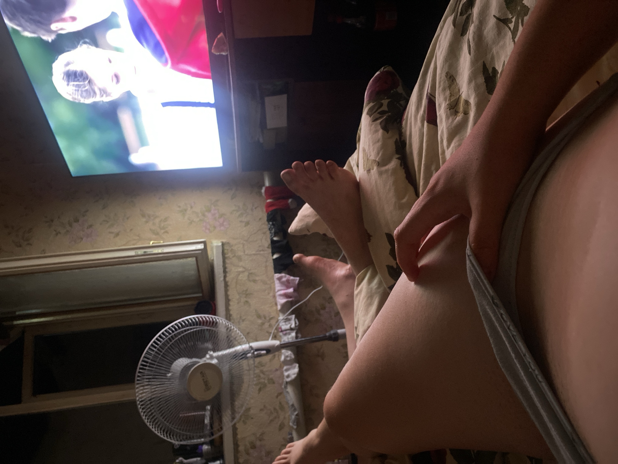 The image shows a close-up view of two people in what appears to be a bedroom or living room. One person's legs and arm are visible, resting on a patterned blanket. They are wearing gray underwear. Their hand is resting on their thigh. The other person's feet are visible in the background, near a fan. A television screen is also visible, displaying an image of a person's face and upper body, with the background blurred and illuminated. The overall lighting suggests it might be evening or nighttime, as the television is the primary light source. The scene conveys a sense of relaxation and casualness, possibly a moment of watching television together. There is no visible text in the image.