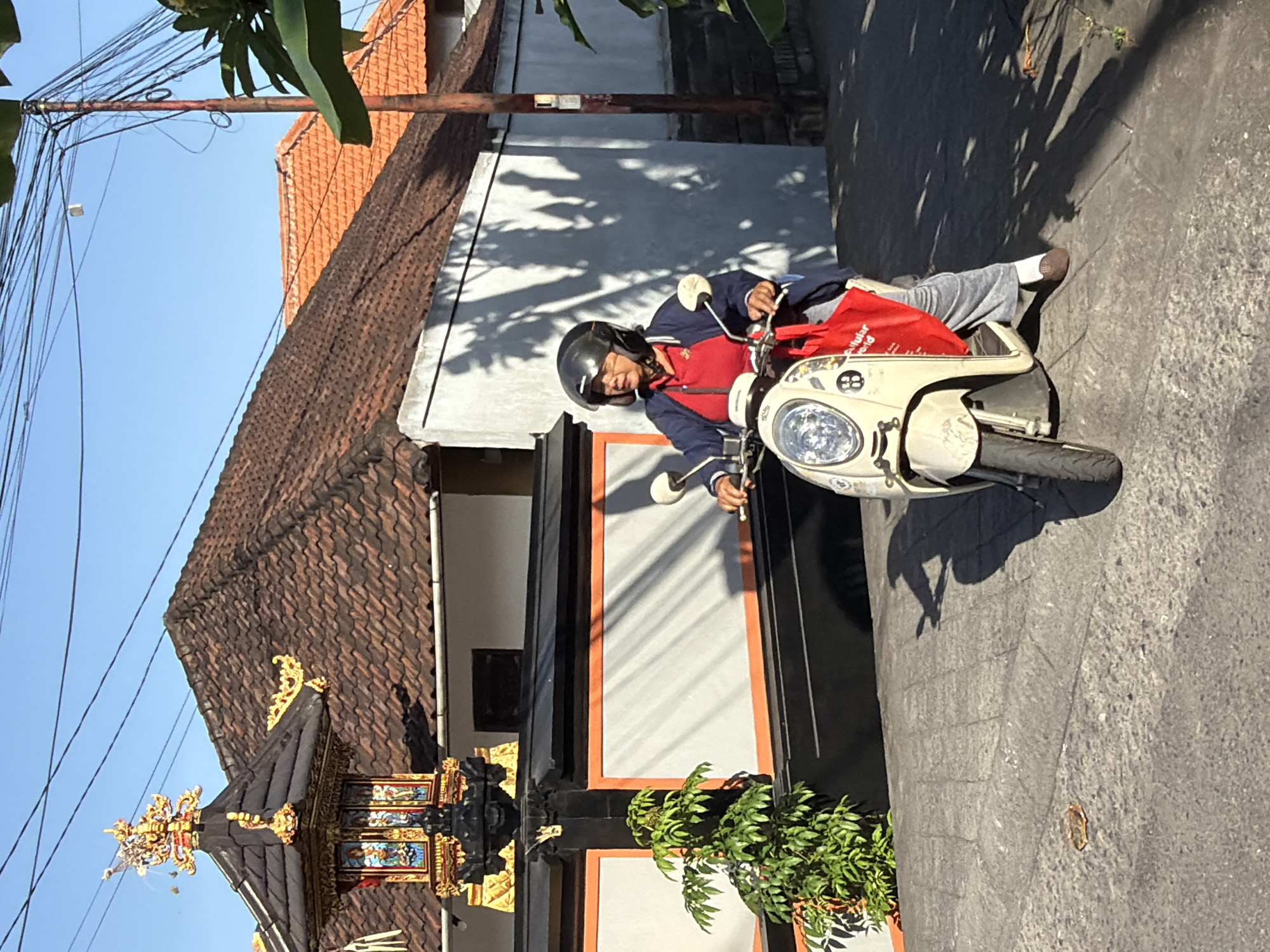 A woman wearing a dark blue jacket over a red shirt and grey pants, with a black helmet, rides a light-colored scooter. She carries a bright red bag on the front of the scooter. The scooter is on a paved road in a residential area. In the background, there are buildings with traditional tiled roofs and a decorative shrine. The scene is illuminated by bright sunlight, casting strong shadows. The location is identified as North Kuta, Indonesia.