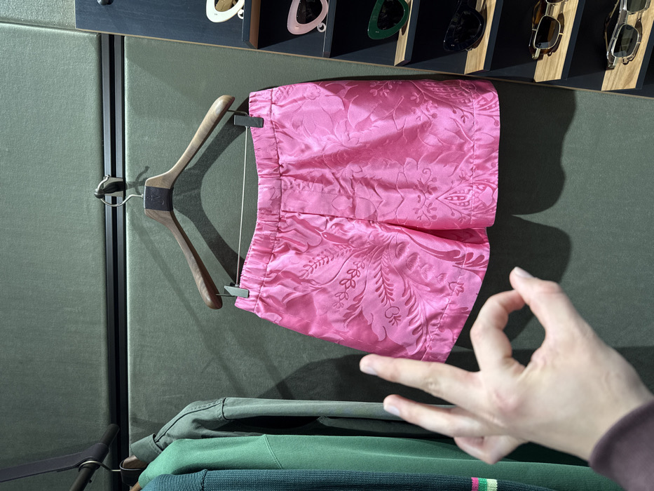 This image captures an interior scene within a retail store, identified as likely a boutique in Paris, France. The central subject is a pair of bright pink shorts, featuring a visible jacquard or embossed floral and paisley pattern, suspended from a dark wooden hanger equipped with metal clips. The shorts display an elasticized waistband. To the left, a dark green knit garment with a striped cuff and a lighter olive-green jacket or shirt are seen hanging on similar wooden hangers. These garments are set against a dark olive-green wall panel, which appears padded or upholstered, segmented by a horizontal dark trim. To the right, a dark wooden shelving unit showcases six pairs of sunglasses. From top to bottom, these include two pairs of white cat-eye frames, one pair of dark green cat-eye frames, one pair of black rectangular frames, one pair of brown tortoise-shell frames with metallic components, and one pair of silver-toned metallic frames. In the lower-left foreground, a left hand, displaying fair skin, performs an 'OK' gesture with the thumb and index finger forming a circle and the remaining three fingers extended. A dark sleeve is partially visible at the wrist.