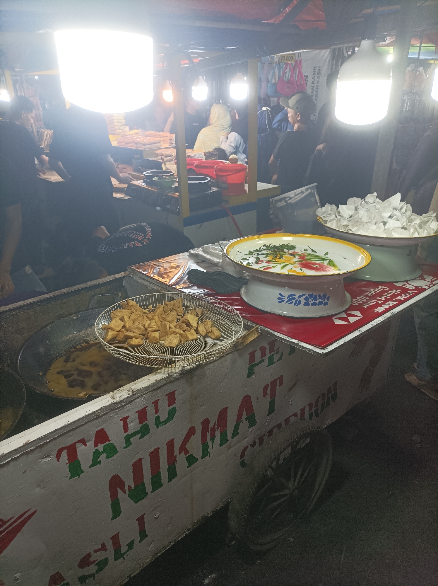 The image shows a food cart at a night market in Cirebon, Indonesia. The cart is brightly lit, with a large, overhead light illuminating the scene. In the foreground, a large wok filled with oil is used for frying tofu. A metal basket containing fried tofu is placed on the cart. Next to the wok, there are two decorative bowls; one contains a green garnish, and the other appears to hold ice. The side of the cart is painted with the words "TAHU NIKMAT" and "ASLI CIREBON" in red and green letters. Several people are visible in the background, some of whom appear to be customers or vendors. The overall atmosphere suggests a bustling street food environment.