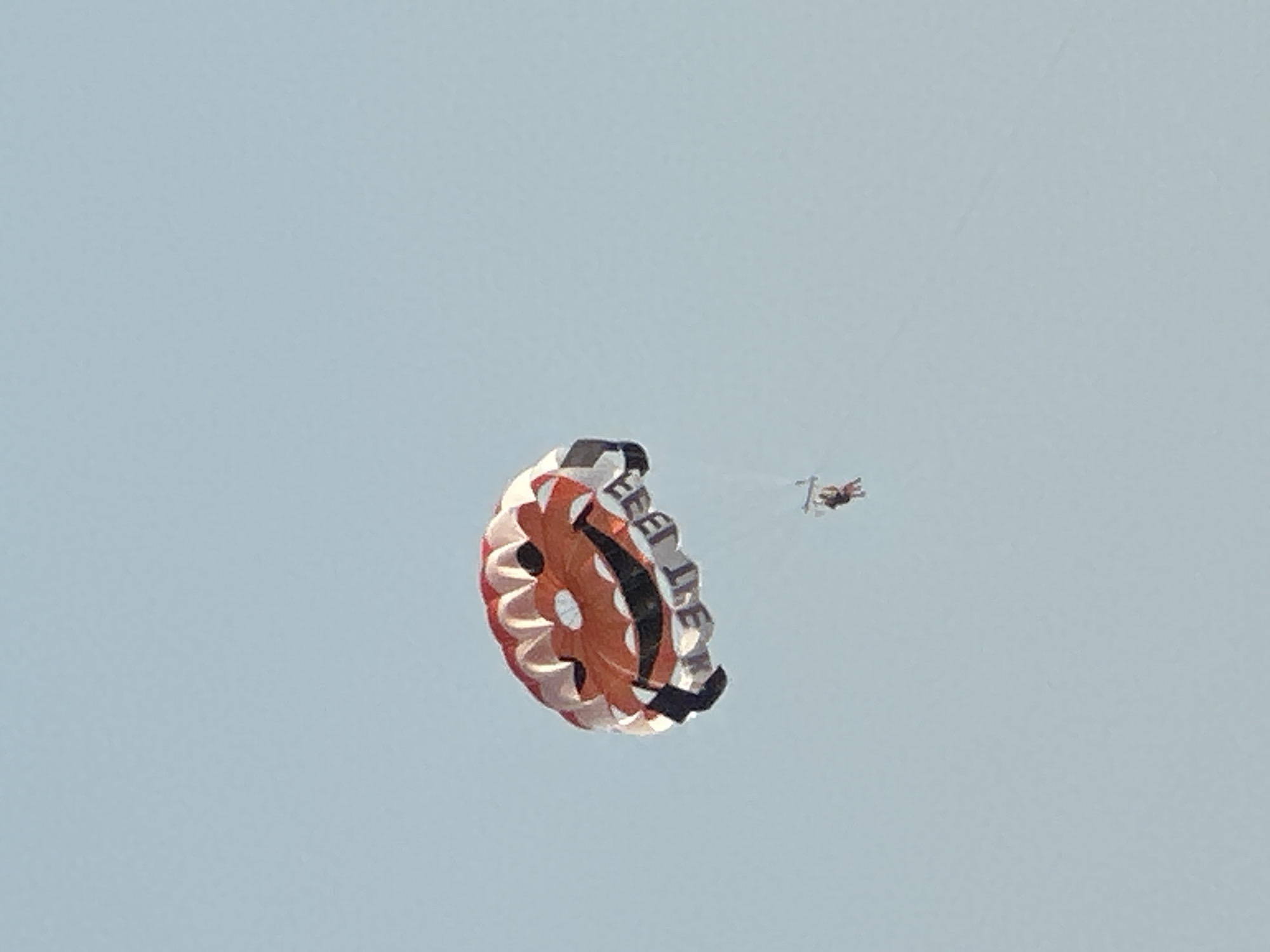 A brightly colored parachute, designed to resemble a smiling face and featuring the text "FEEL IT" is suspended in a clear, pale blue sky. Below the parachute, two figures are harnessed and dangling from tow lines, engaged in parasailing. The parachute is predominantly orange and white with black accents forming the smiley face. The parasailers are small in the frame, appearing as dark silhouettes against the sky, suggesting they are in mid-air, likely over the water, given the context of parasailing. The scene is devoid of any identifiable urban or natural landmarks that would place it specifically within Boreti, Montenegro, as the image focuses solely on the sky and the activity occurring within it.