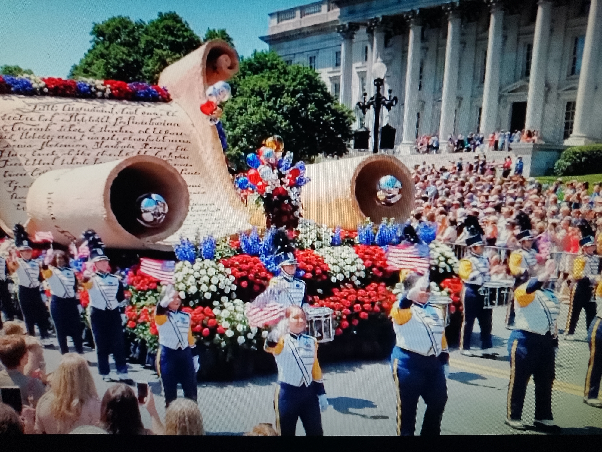 The image depicts a parade featuring a large scroll-shaped float adorned with red, white, and blue flowers. A marching band in uniform, consisting of individuals in navy pants, white tops with yellow accents, and black hats, is visible in front of the float. Some band members are playing instruments or saluting. In the background, a crowd of spectators is gathered, and a grand building with columns, possibly a government building or monument, stands behind them. The scene appears to be a patriotic celebration. There is no indication that this image is located in Chiajna, Romania; the architecture and the presence of a marching band in such a setting are not indicative of that specific location.