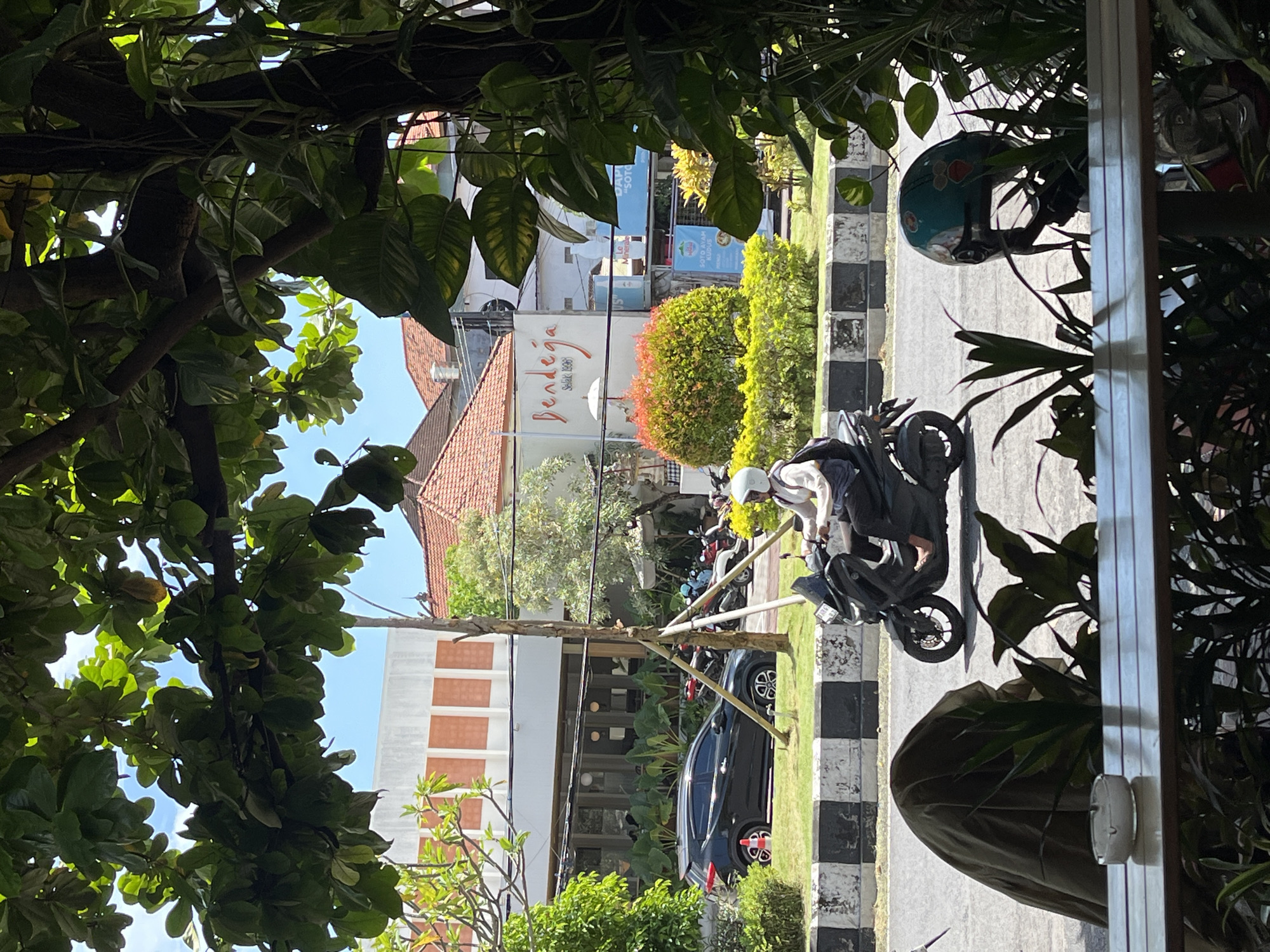 The image shows a street view in Kuta Selatan, Indonesia. A person on a black motorbike with a white helmet is stopped along a curb with black and white markings. The background features buildings, including one with the word "Bendega" written on it. A black car is parked nearby. The scene is framed by foliage in the foreground.
