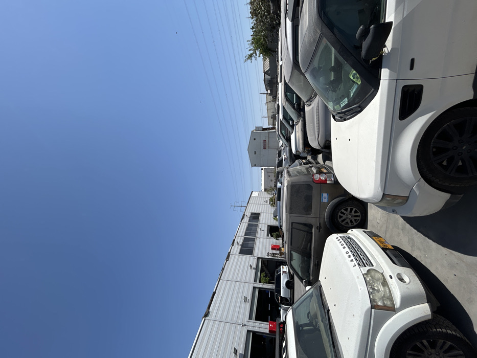 Rows of Land Rover SUVs fill a parking lot beside a large, white industrial building. The vehicles are a mix of white, gray, and tan colors. A clear blue sky is overhead, with power lines visible. The image is rotated to a 90-degree angle. One SUV in the center has a visible flag on the rear tire.