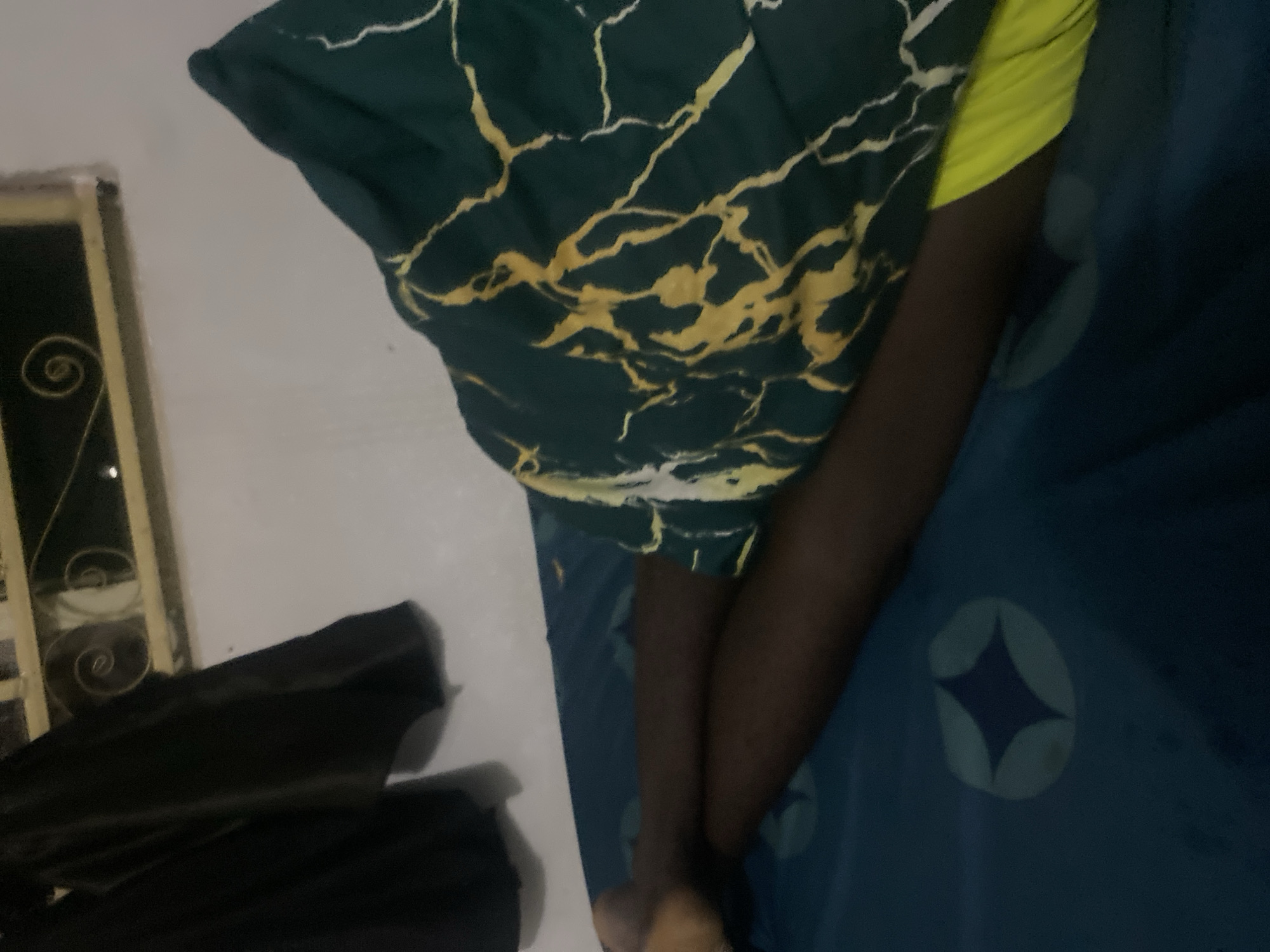 The image depicts a person lying in bed, partially covered by a dark green blanket with a yellow marble-like pattern. The person's legs, wearing bright yellow shorts, are visible beneath the blanket. The bed has a blue sheet with a star-like pattern. In the background, a black garment hangs on the wall, and a window with decorative metalwork is visible. The setting appears to be a bedroom in Howayi, Nigeria.