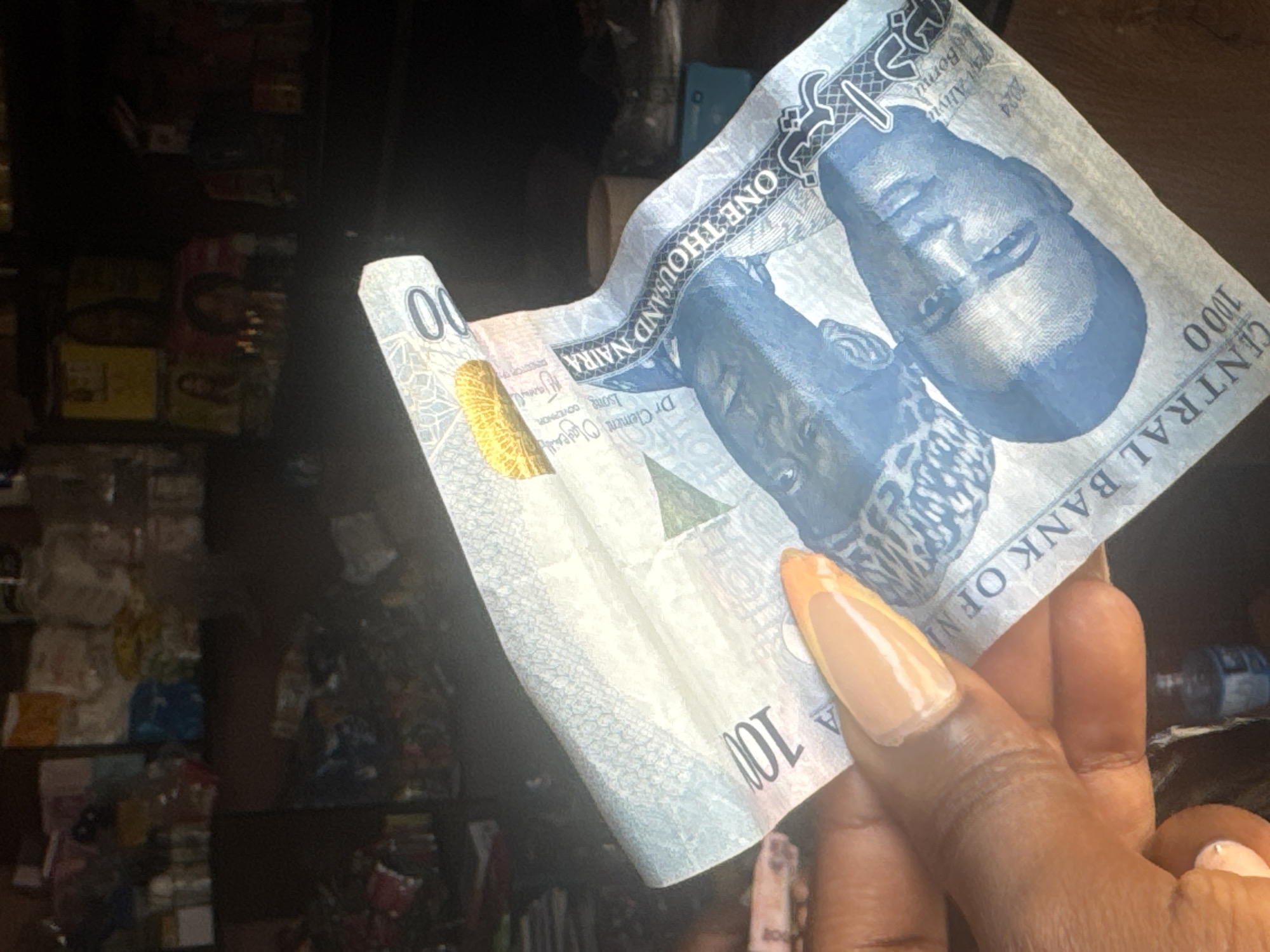 The image shows a close-up of a hand holding a Nigerian 1000 Naira banknote. The banknote is bent and appears to be made of polymer. The hand has long, manicured nails with a French tip design. The banknote features portraits of Nigerian leaders and text, including "CENTRAL BANK OF NIGERIA," "ONE THOUSAND NAIRA," and the year "2024." The background is dark and blurry, with hints of shelves filled with various items, suggesting a retail or market environment. The lighting appears to be artificial, possibly from indoor lighting. The overall impression is a transaction or a display of currency.