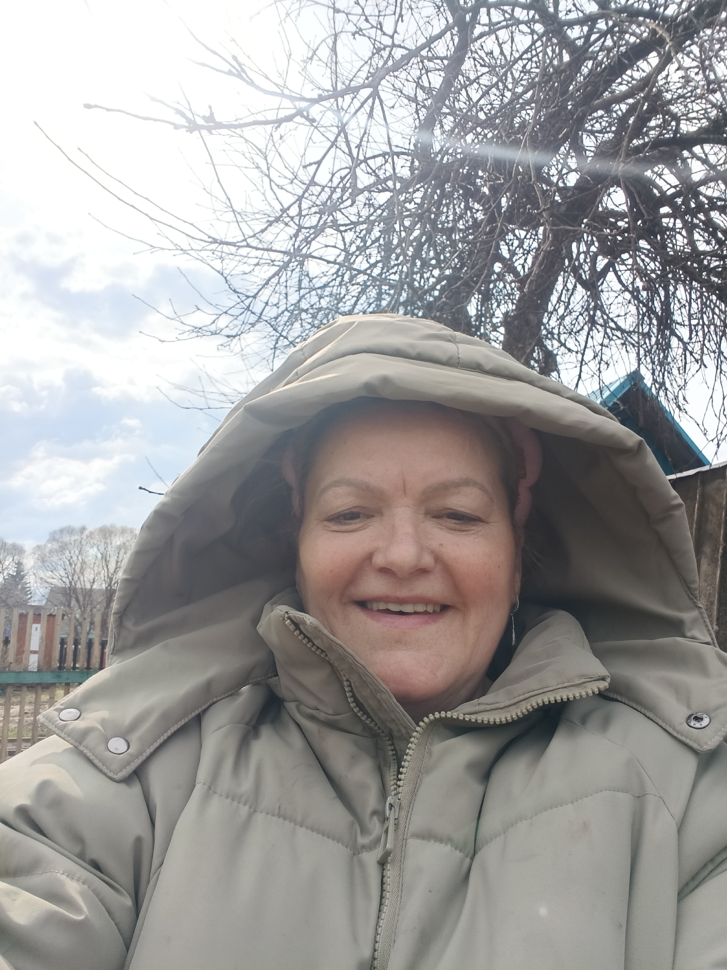 A smiling, middle-aged to older woman is featured in a bright outdoor selfie, likely captured in Krasny Mayak, Russia. She faces the camera directly, displaying a broad, warm smile that conveys happiness or contentment. She is dressed in a light beige or khaki puffer jacket with the hood pulled up over her head, covering much of her hair, though a glimpse of a pink earmuff or headband is visible. A silver-toned dangle earring can be seen on her left ear.
The setting is outdoors, suggesting a rural or village environment during late winter or early spring. Behind her, a dense network of bare tree branches, indicative of a deciduous tree, dominates the upper and right portions of the background, silhouetted against a bright, cloudy sky. The sun, positioned in the upper left, creates a strong backlight and a lens flare, washing out that section of the sky.
To the left of the woman, a rustic wooden fence is visible, beyond which stand distant trees and what appear to be other structures under a partly cloudy, light blue sky. On the far right, a corner of a building with a distinctive turquoise roof and a brown wall or fence is partially visible through the bare branches. The image captures a casual, cheerful moment in a bright, cool weather setting. No visible text is present.