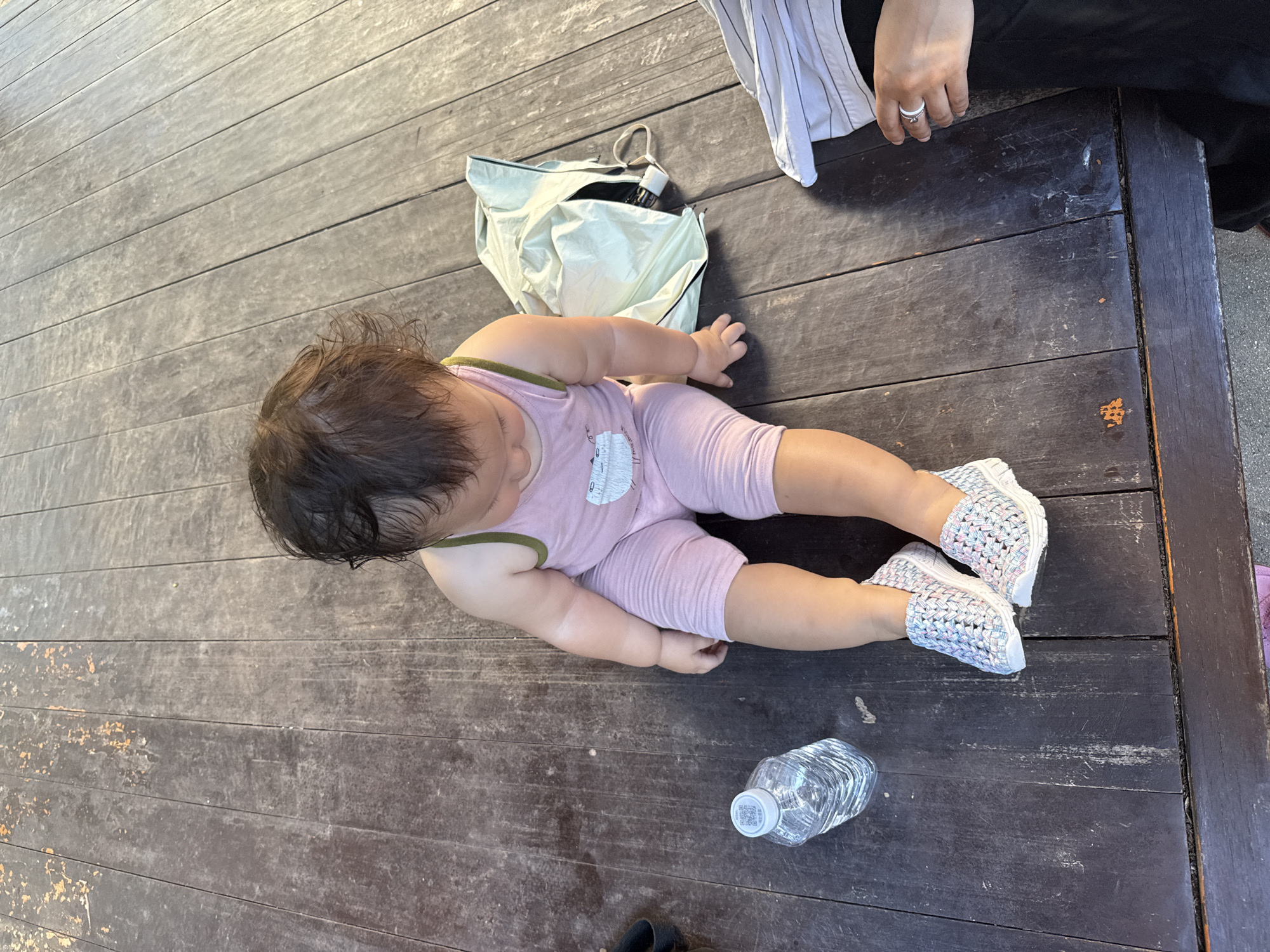 An overhead, medium shot captures a young child with dark hair sitting on a weathered, dark wooden deck. The child is wearing a light pink tank top with a white graphic and light pink leggings, along with white and multi-colored woven slip-on shoes. To the left of the child is a clear plastic water bottle with a white cap. Behind the child is a light green drawstring bag, partially unzipped, with an object inside. To the right of the frame, a person's hands are visible, wearing a ring on their left ring finger, and their black clothing. The wooden deck shows signs of wear, with some areas of faded paint or discoloration. The lighting suggests it is daytime, with shadows cast on the deck. The image was taken in Gwangju, South Korea.