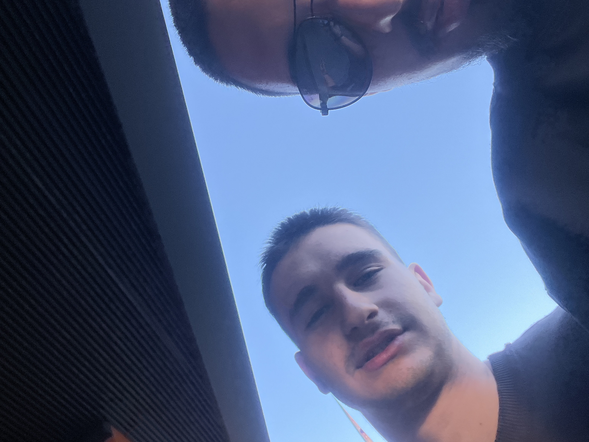 A selfie taken from a low angle shows two men under a bright, blue sky. The man in the foreground is facing the camera, with a closed right eye and a slightly open mouth. The man behind him is wearing sunglasses. The location includes a modern building with horizontal lines, suggesting an urban environment. Although there are no clear indicators to confirm its location in Plovdiv, Bulgaria, the environment is consistent with an urban setting.