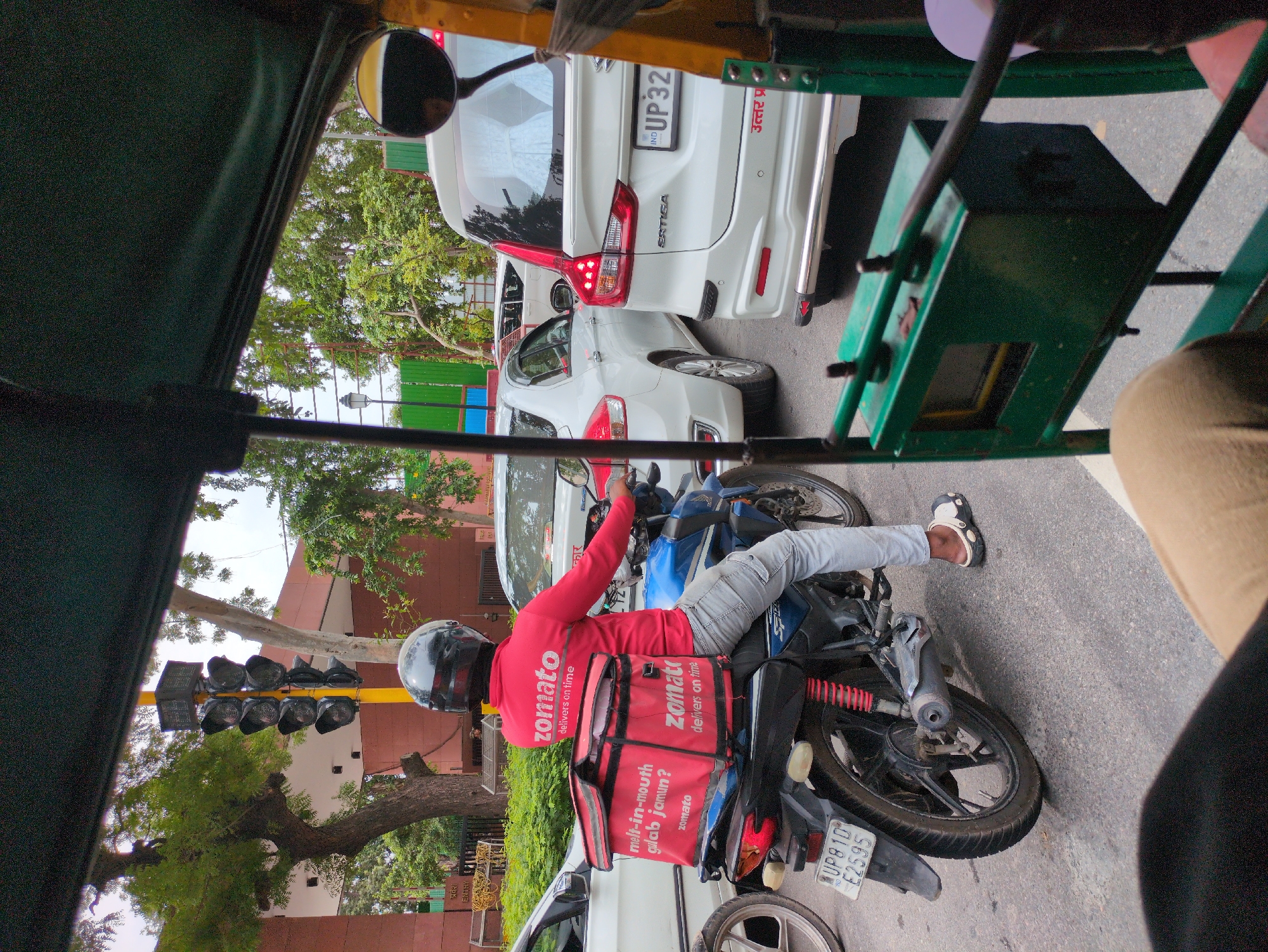The image captures a street scene in New Delhi, India, likely during daylight hours with overcast skies. The perspective is from inside an auto-rickshaw, looking forward and slightly to the right.
In the foreground, the front of the auto-rickshaw is visible, with a green metal frame and some controls. A person's leg, wearing a light brown legging and a beige and white sandal, is visible in the bottom right.
The main subject is a Zomato delivery person on a blue and black motorcycle, stopped in traffic. The rider is wearing a red full-sleeved jacket with the Zomato logo and "delivers on time" printed in white. They have a black helmet on and are wearing grey trousers. A large red Zomato delivery bag is strapped to the back of the motorcycle. Text on the bag includes "melt-in-mouth gulab jamun?" and the Zomato logo. The motorcycle has a license plate visible: "UP81D E2595".
Behind the motorcycle, there is a significant traffic jam of white cars. The closest car to the motorcycle is a white sedan. Further back, a white SUV (an Ertiga) is visible with a license plate that starts with "IND UP32". Red lettering, possibly Hindi text, is also visible on the rear bumper of the SUV.
In the background, trees and foliage line the street. A traffic light signal with multiple lamps is mounted on a yellow pole. A building with red brick walls and white accents is visible to the left, with trees in front of it. A metal fence is also present in the background. The overall environment suggests a busy urban area.