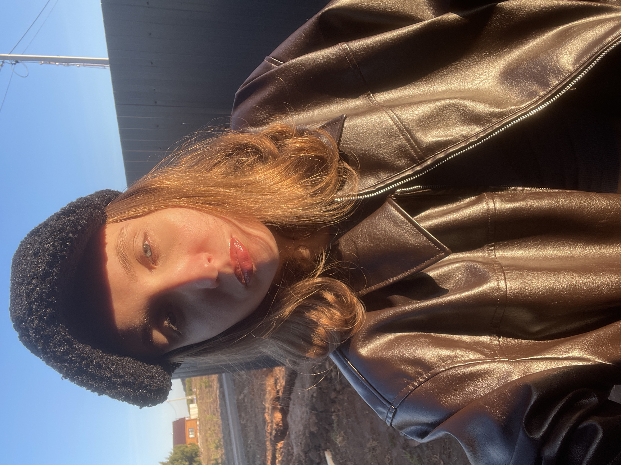 The original image, which is rotated 90 degrees counter-clockwise, depicts a young person outdoors under strong, direct sunlight.
The central subject is a young individual, likely female, positioned slightly off-center to the left. They have light brown or auburn hair, visible framing their face and cascading over their shoulder. Their eyes are light-colored, appearing green or hazel. They wear a dark, textured hat, possibly a beanie or bucket hat, and glossy, reddish lip product is visible on their lips. The individual is dressed in a shiny, dark brown faux leather jacket, featuring visible seams and a silver zipper that is partially unzipped.
The background is an outdoor environment. The upper left portion of the frame shows a clear, bright blue sky with a utility pole or thin structure and wires against it. The lower right background reveals dry, light-colored ground or sparse vegetation, with indistinct structures, possibly a building, in the distance. A dark, corrugated structure, possibly a fence or wall, is visible behind the person's head, separating the sky from the ground.
The image captures a posed moment, likely a selfie or portrait, with the person looking towards the camera. Strong, direct sunlight illuminates the scene, casting prominent highlights on the person's hair and the glossy texture of the jacket. A significant shadow falls across the left side of the person's face, partially obscuring their eye and cheek, creating a dramatic contrast. The intense light and warm tones suggest the photo was taken during the golden hour, either early morning or late afternoon, on a clear, sunny day. No text is visible in the image. The location context for this scene is Podshivalovo, Russia.