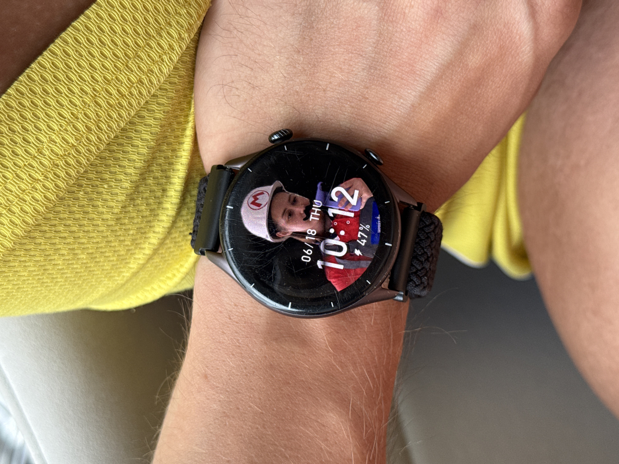 An image shows a close-up of a person's wrist wearing a smartwatch. The watch face displays the date as 06/18 THU and the time as 10:12. A picture is displayed on the watch face, featuring a person wearing a white hard hat with a red "M" logo and a fake mustache. The watch also shows the battery level at 47%. The person is wearing a bright yellow shirt. The background appears to be indoors with a muted, possibly beige, surface and some light. The image does not contain any information or visual cues that would allow for the determination of the location in Chaikovsky, Russia.