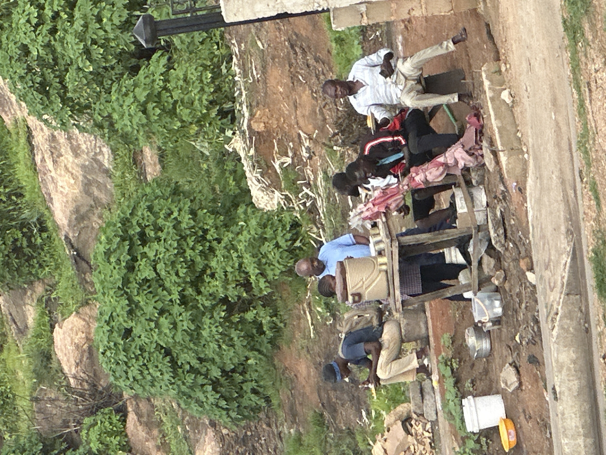 The image depicts an outdoor scene in Gura Top, Nigeria, where several individuals are gathered around a makeshift food stall. In the foreground, a young man wearing a blue t-shirt and light-colored trousers sits on a low concrete structure, eating from a plate. He has a backpack slung over his shoulders. To his right, a food stall with a wooden top displays various items, including what appears to be a large beige food container and metal bowls. An older man in a light blue shirt stands behind the stall, looking towards the right. Further to the right, another man in a light-colored striped shirt and light trousers sits on a bench, holding a plate and a piece of food. To his left, two other men are seated, with one wearing a red and black jacket. The background shows a rocky, uneven terrain with lush green vegetation and some trees. The ground around the stall is dirt and debris. A white bucket and an orange bowl are visible on the ground near the left side of the image. A metal railing and a dark pillar are present on the far right edge of the frame. The overall atmosphere suggests a casual gathering or a roadside food service.