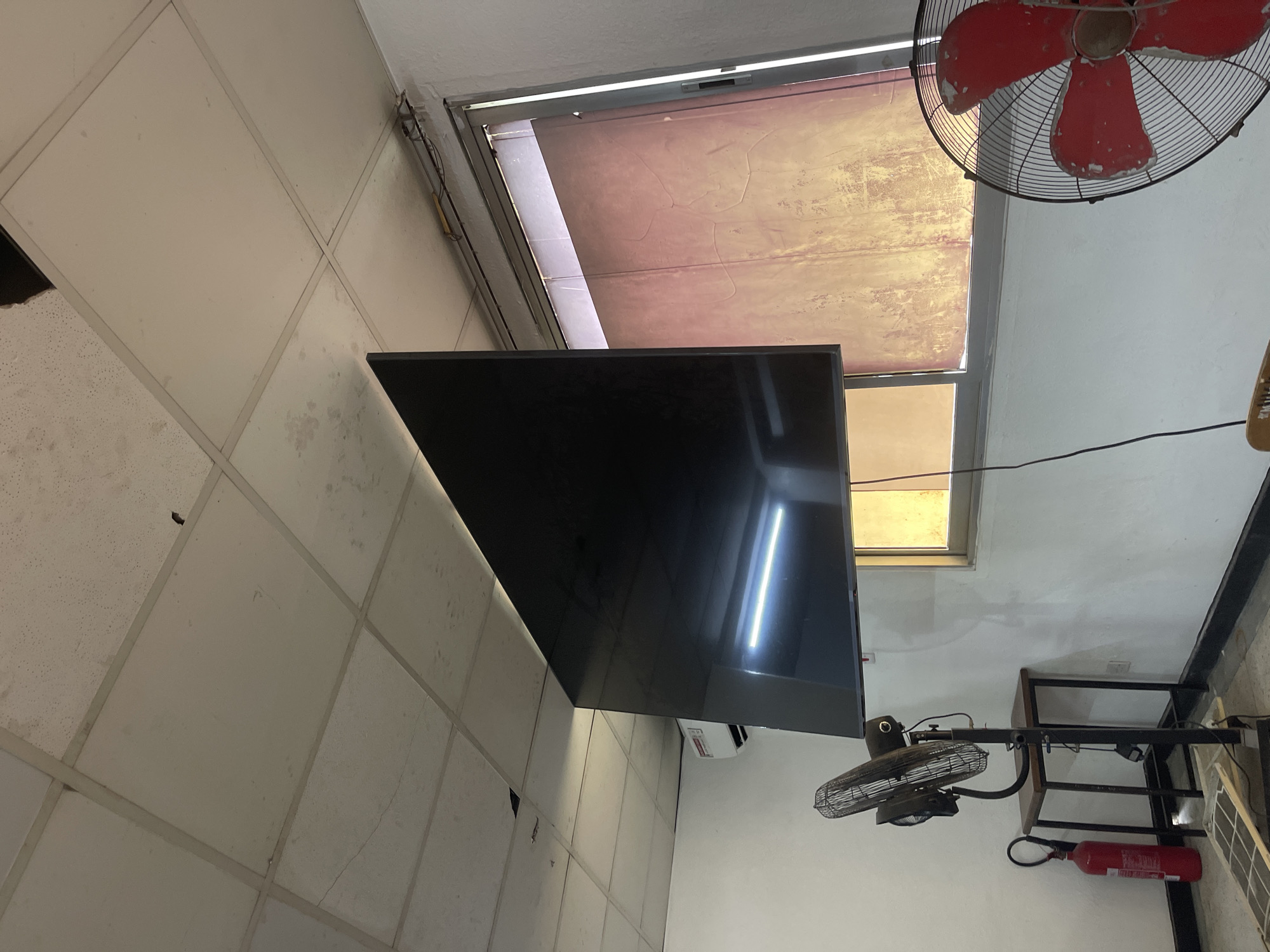 The image captures an indoor scene, likely a commercial or public space, in Dar es-Salaam, Tanzania, taken from a slightly upward, tilted angle. The setting features a drop ceiling composed of white acoustic tiles, many of which exhibit significant discoloration, stains, and signs of wear, suggesting age or potential moisture issues.
A prominent object is a large, black flat-screen television mounted high on a wall, reflecting a bright, vertical light source. Below and to the right of the television, a small white air conditioning unit is partially visible. Two electric fans are present: a large, red, industrial-style fan with a protective cage, possibly ceiling or high-wall mounted, showing chipped paint on its blades, and a smaller, black stand fan located lower on the right side of the frame.
A window is visible in the background, covered by a light-colored, possibly translucent or aged, blind or film that diffuses natural light into the room. The window frame appears metallic. Below the smaller fan, a red fire extinguisher stands on the floor or a low surface, though any text on it is unreadable. A simple black metal-framed table or shelving unit is also visible. Black electrical wires run along the walls, connecting various appliances.
No people are present, and no specific activity is taking place, but the arrangement of a TV, fans, and AC suggests a space designed for comfort and information display. The scene appears to be set during the daytime due to the natural light from the window, supplemented by indoor lighting as evidenced by the reflection on the TV. The overall impression is that of a functional, well-used space showing signs of age and wear.