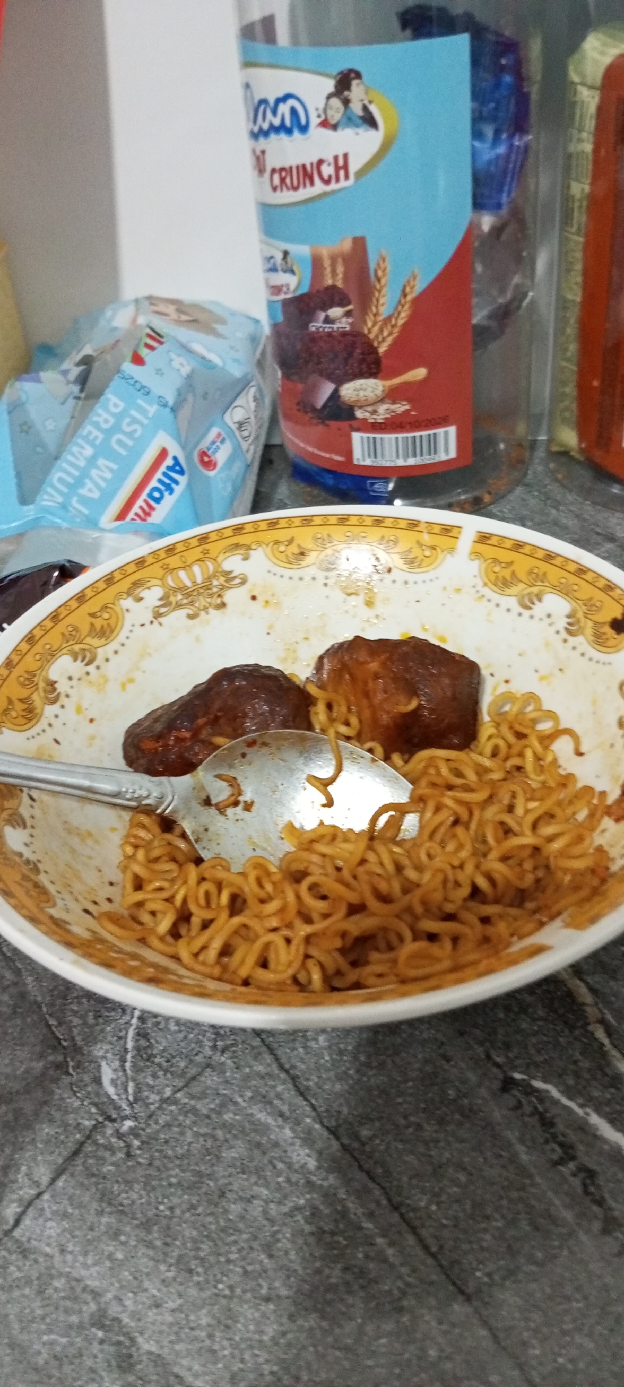 A close-up, eye-level shot captures a bowl of instant noodles with two pieces of meat, likely chicken, and a spoon. The noodles are golden brown and appear to be sauced, while the meat is dark brown and textured. The bowl is white with a decorative gold rim. In the background, a blue and white box labeled "Alfamart" is visible, along with a plastic container with a label that includes the words "DANCOW CRUNCH" and images of a chocolate bar and wheat. The setting appears to be an indoor environment, possibly a home kitchen or dining area, with a gray, marbled surface in the foreground. The lighting is bright, suggesting natural or artificial light sources overhead. There are no individuals visible in the image. The photograph's perspective and focus highlight the food and surrounding items on a countertop.