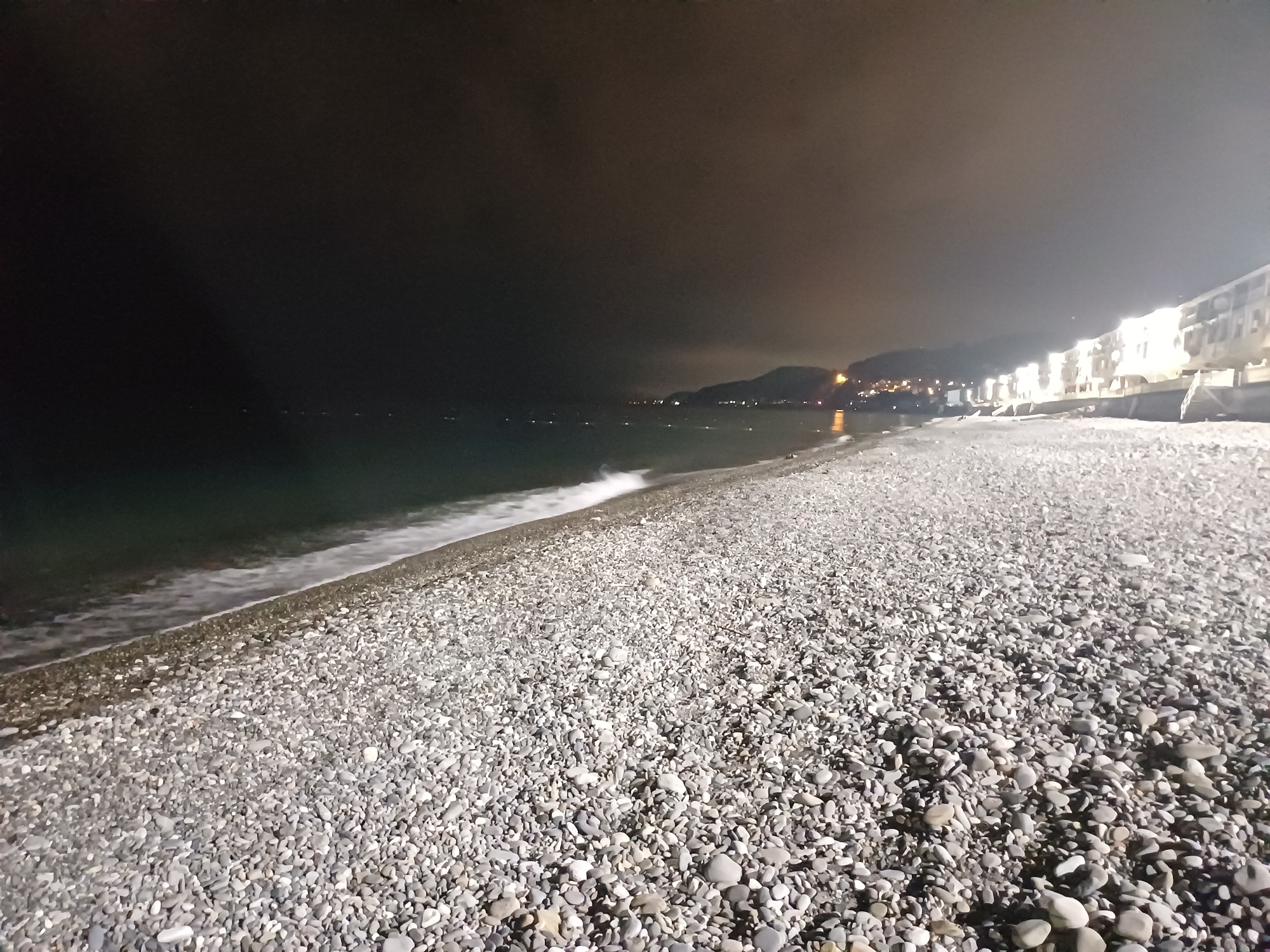 This nighttime image depicts a wide view of a pebble beach in Sochi, Russia. The foreground is dominated by a vast expanse of irregularly shaped grey and white pebbles covering the beach, extending towards the upper right of the frame. To the left, the dark water of the sea meets the shore, with a line of white foam from a breaking wave visible along the edge. Several faint, distant lights are scattered across the surface of the dark sea. In the mid-ground, a coastline with dark, undulating terrain, likely mountains or hills, recedes into the distance. Bright clusters of urban lights are visible along this distant coastline. To the right, a series of multi-story buildings, highly illuminated, stand prominently along the beachfront, casting a strong light. The sky above is dark and overcast, exhibiting a diffused orange-brown glow, indicative of light pollution. No individuals or specific human activities are discernibly present in the scene.