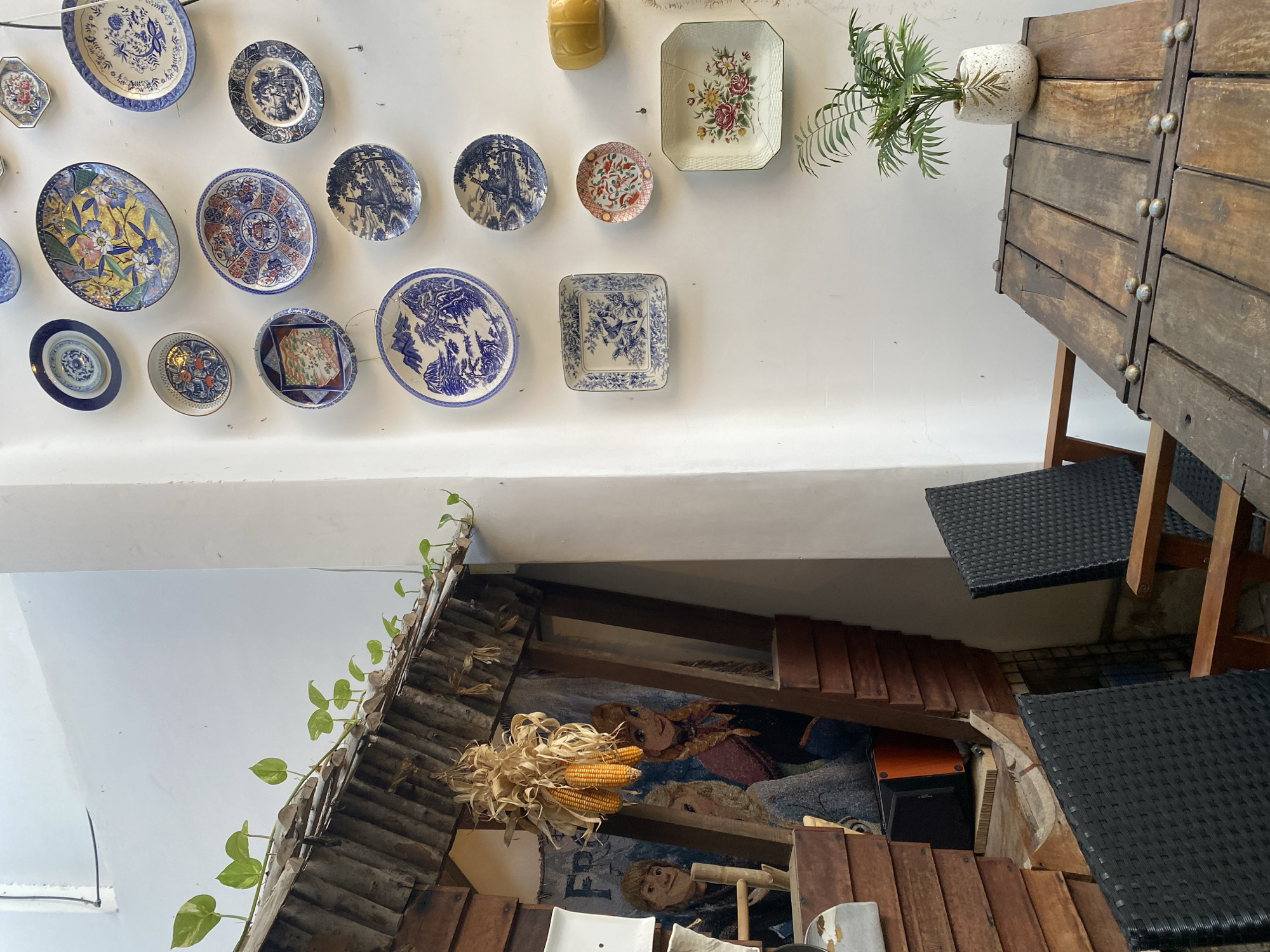 The image depicts an outdoor seating area, likely a cafe or restaurant, featuring a rustic wooden table and chairs against a white wall adorned with decorative plates. Vines trail along the edge of a wooden roof structure on the left. Hanging from the roof are bunches of dried corn cobs. A mural of cartoon characters, possibly from the movie Frozen, is visible behind the corn. The table is set with a potted green plant. The overall setting suggests a charming, somewhat eclectic atmosphere. The location is specified as Nha Trang Ward, Vietnam.